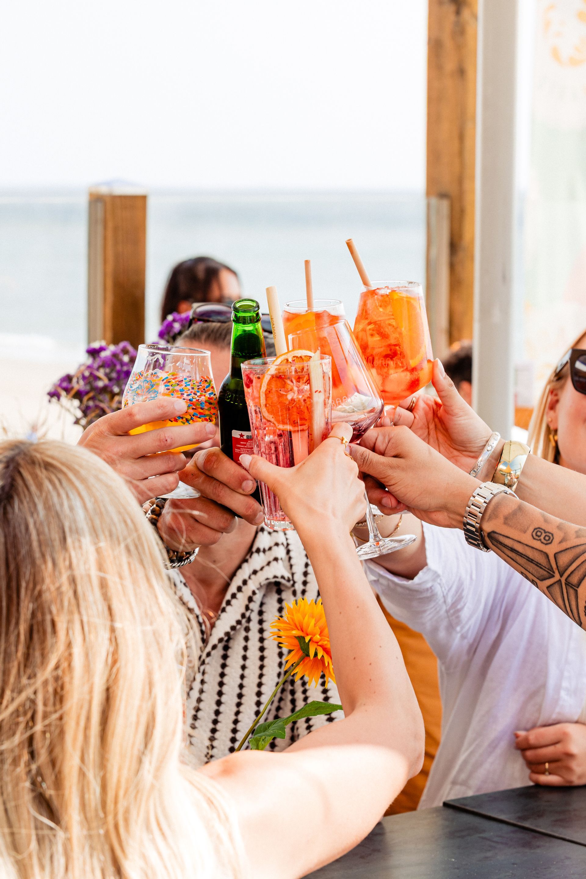 Mensen brengen buiten, vlakbij het water, een toast uit en vieren feest met kleurrijke cocktails.