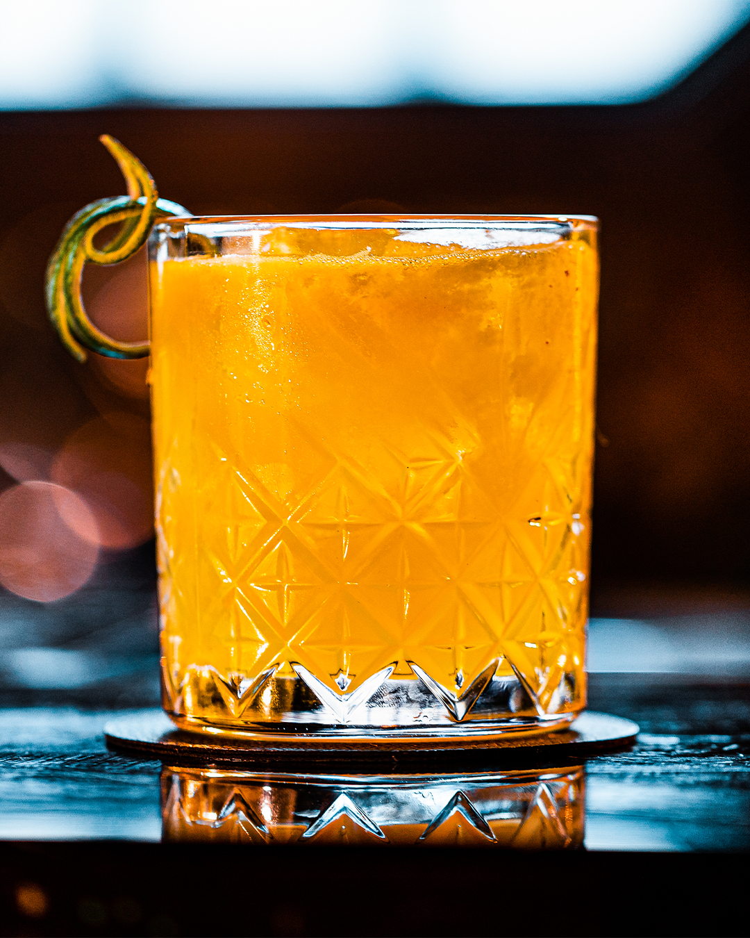 Een sinaasappelcocktail in een glas met patroon, gegarneerd met een schijfje citrusvrucht, staat op een donkere onderzetter.
