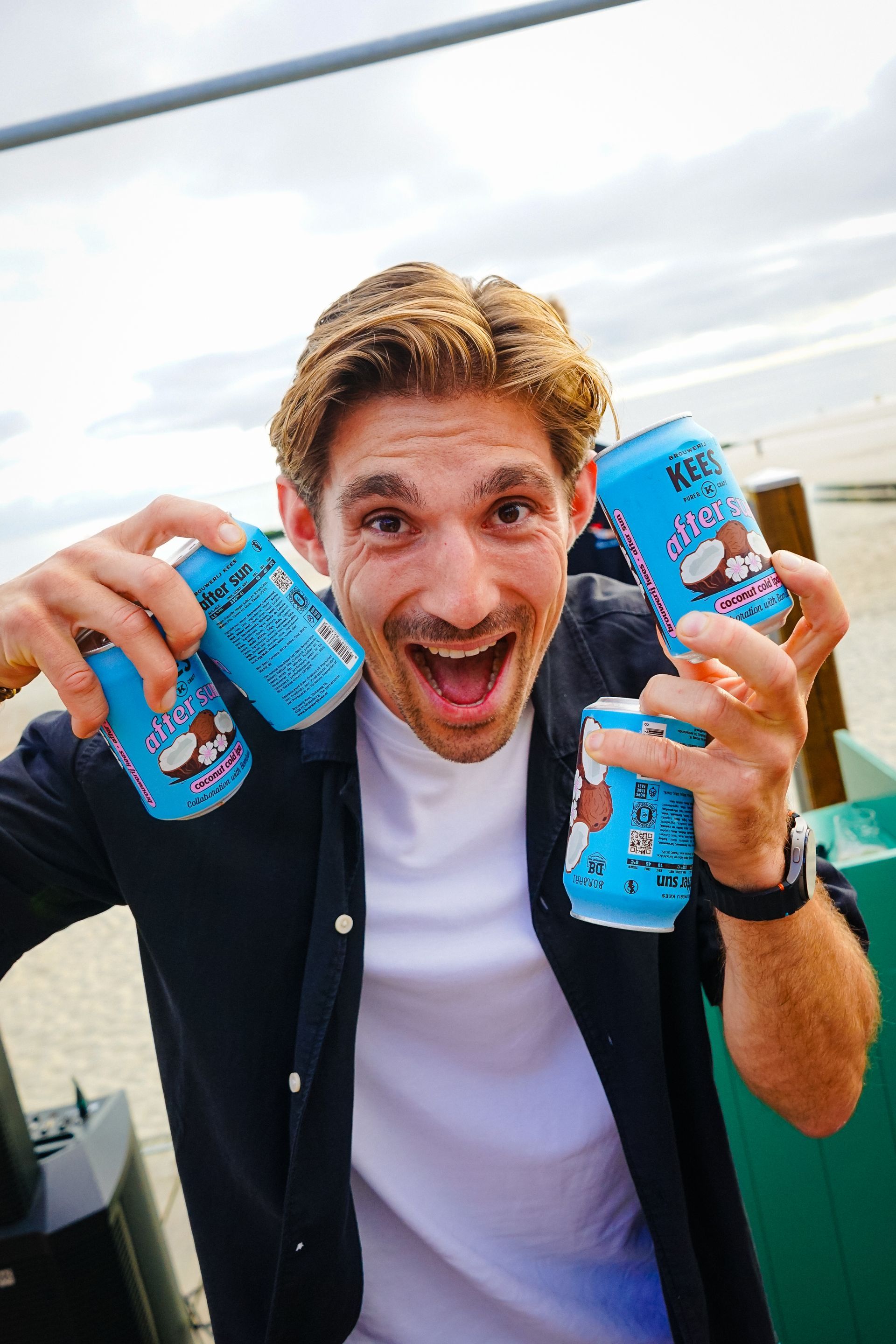 Een man houdt lachend vier blikjes van een lichtblauwe drank met een kokosnootafbeelding vast.