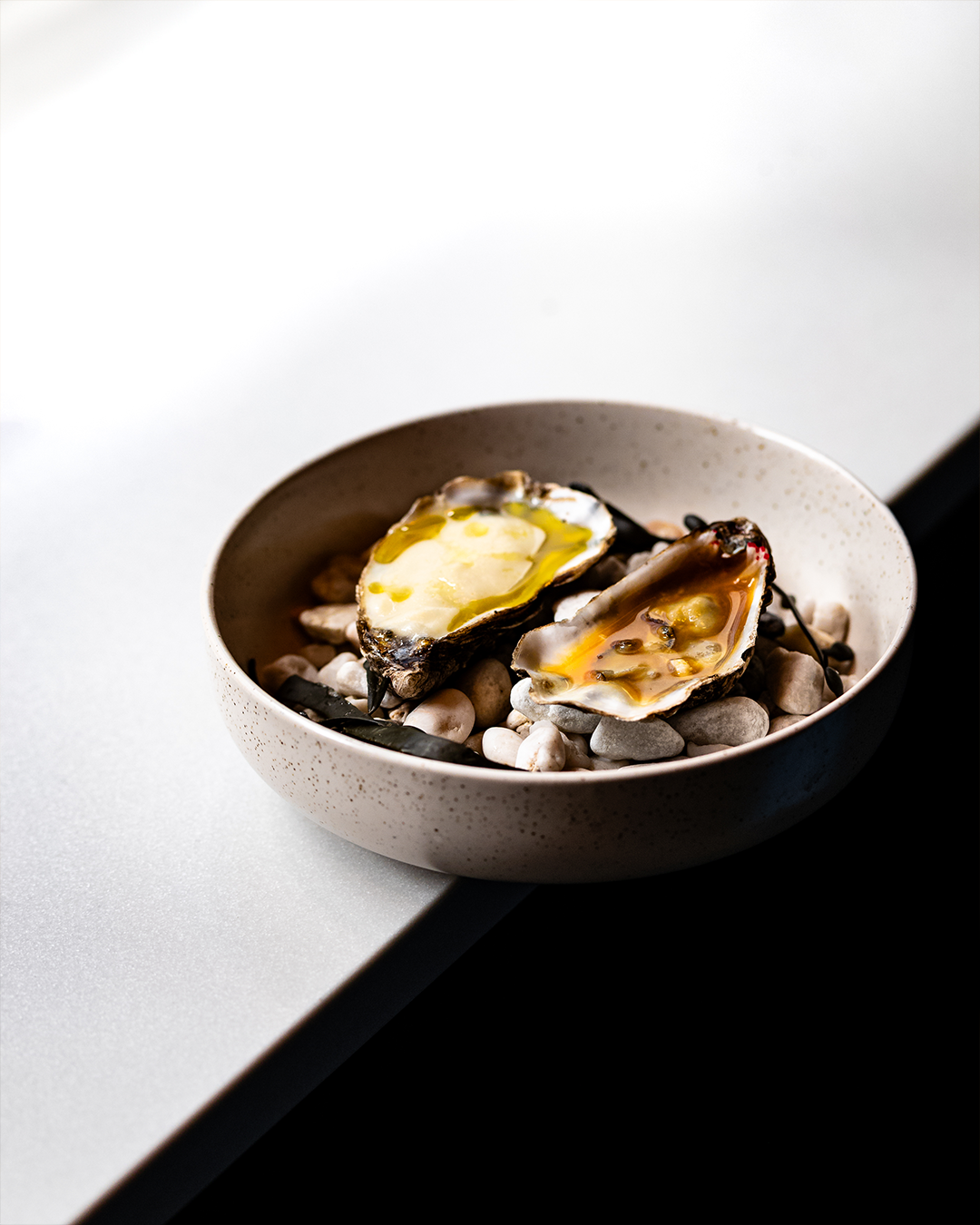 Oesters, gebakken in hun schelp met diverse toppings, geserveerd in een gespikkelde kom op stenen, afstekend tegen een zwart-witte achtergrond.
