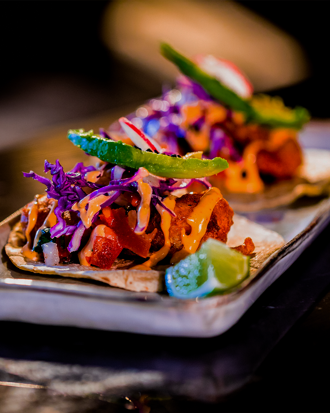 Twee rijkgevulde taco's op een bord, met gefrituurd voedsel, paarse kool, saus en limoen.