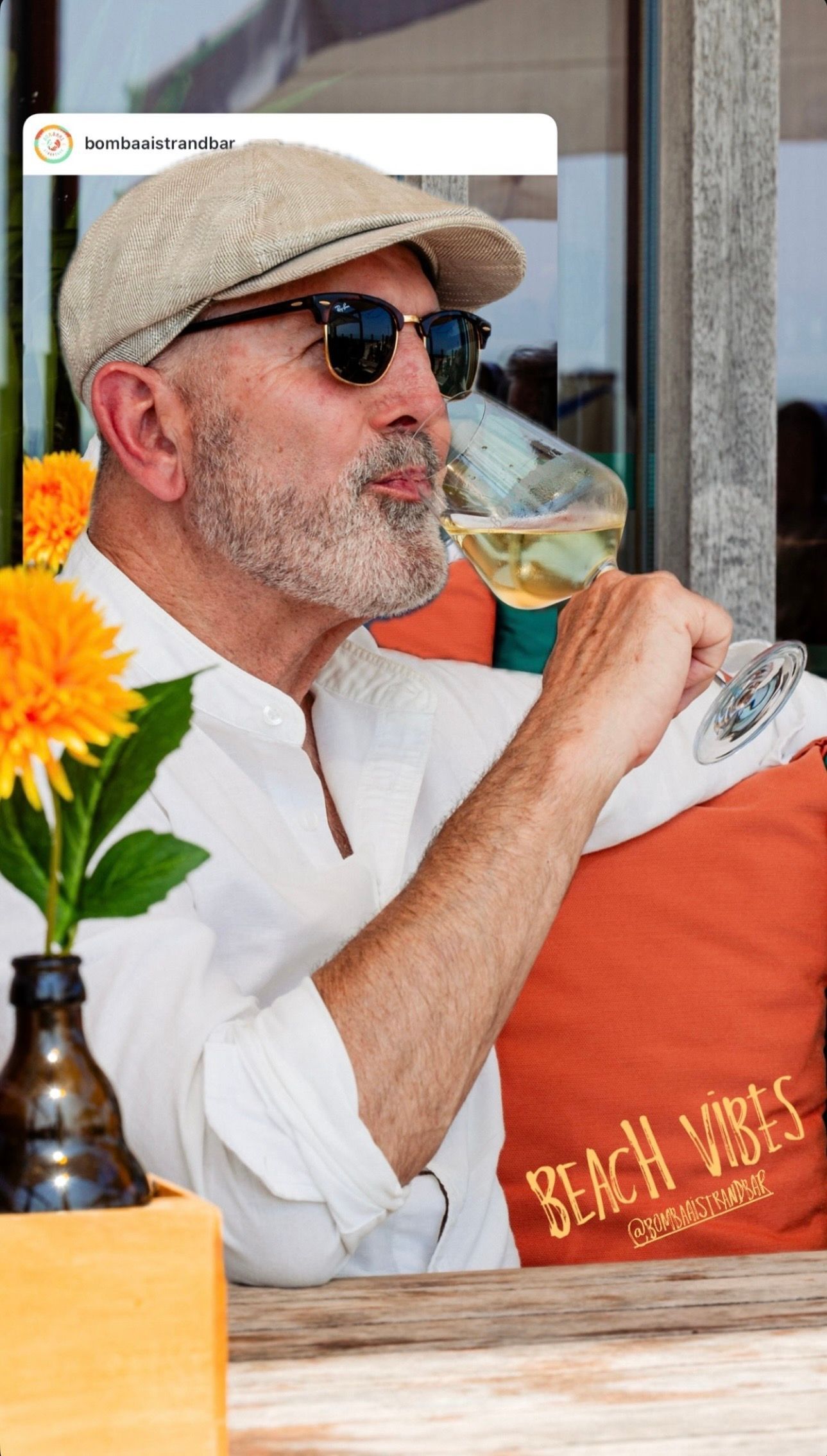 Een man zit buiten aan een glas wijn, draagt een zonnebril en een pet. Op een oranje kussen staat