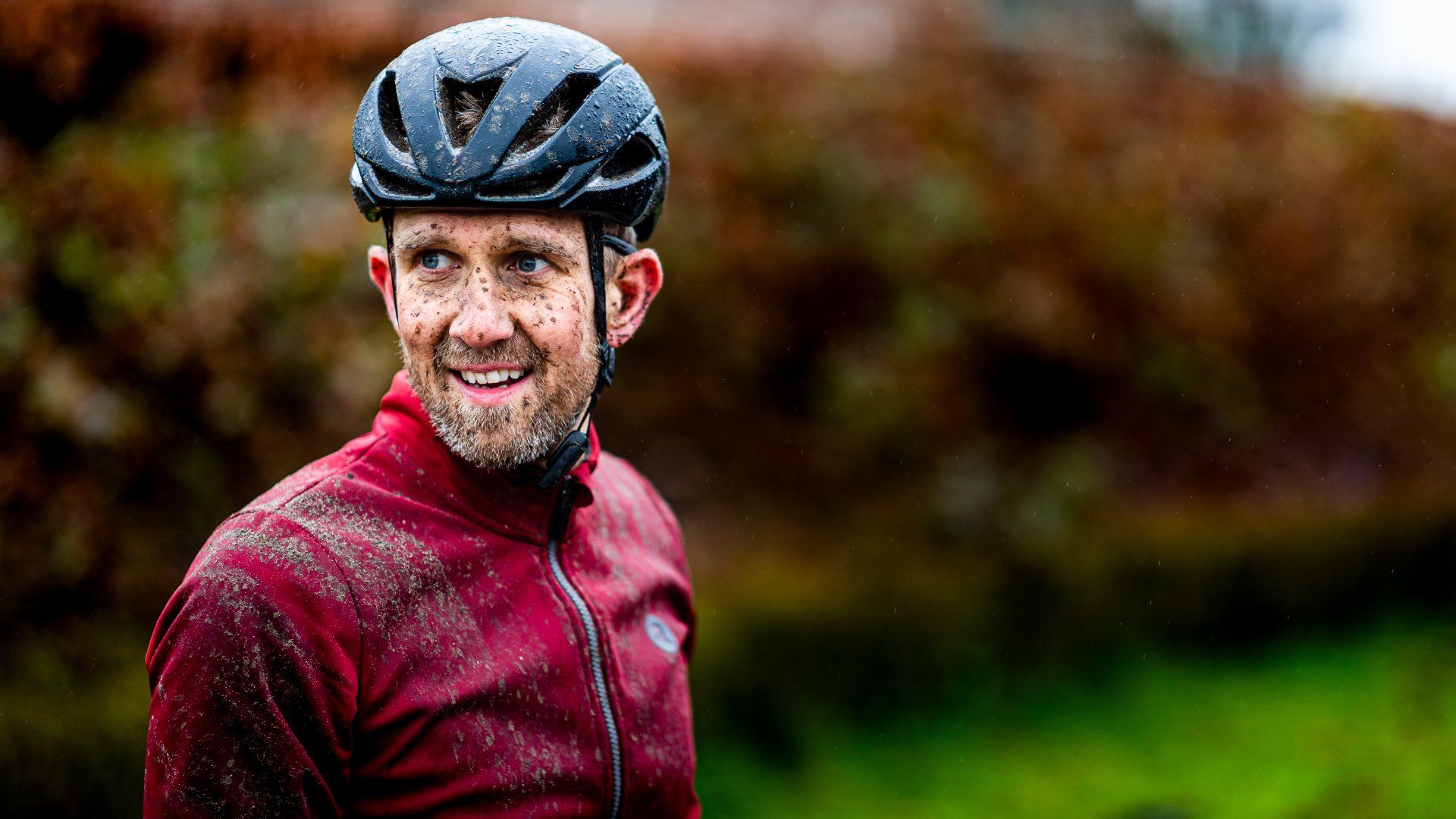 Een man met een fietshelm en een rode jas, onder de modder, staat buiten te lachen.