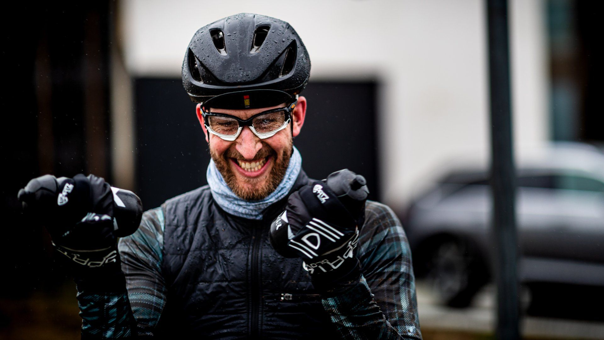 Een fietser met helm, bril en handschoenen lacht en houdt zijn vuisten omhoog; de foto is buiten genomen.
