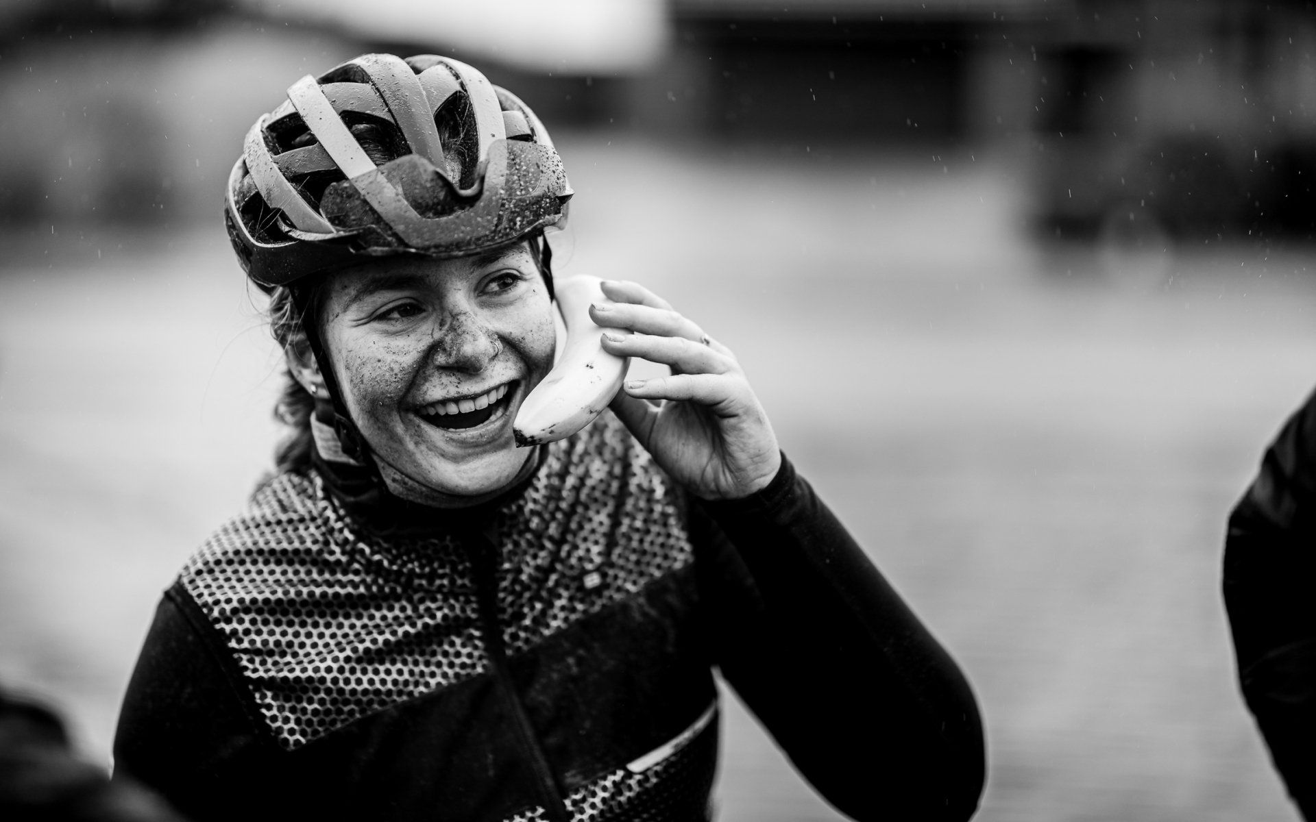 Een lachende fietser houdt een banaan tegen haar oor alsof het een telefoon is. Ze draagt een helm en fietskleding. Buiten, zwart-wit.
