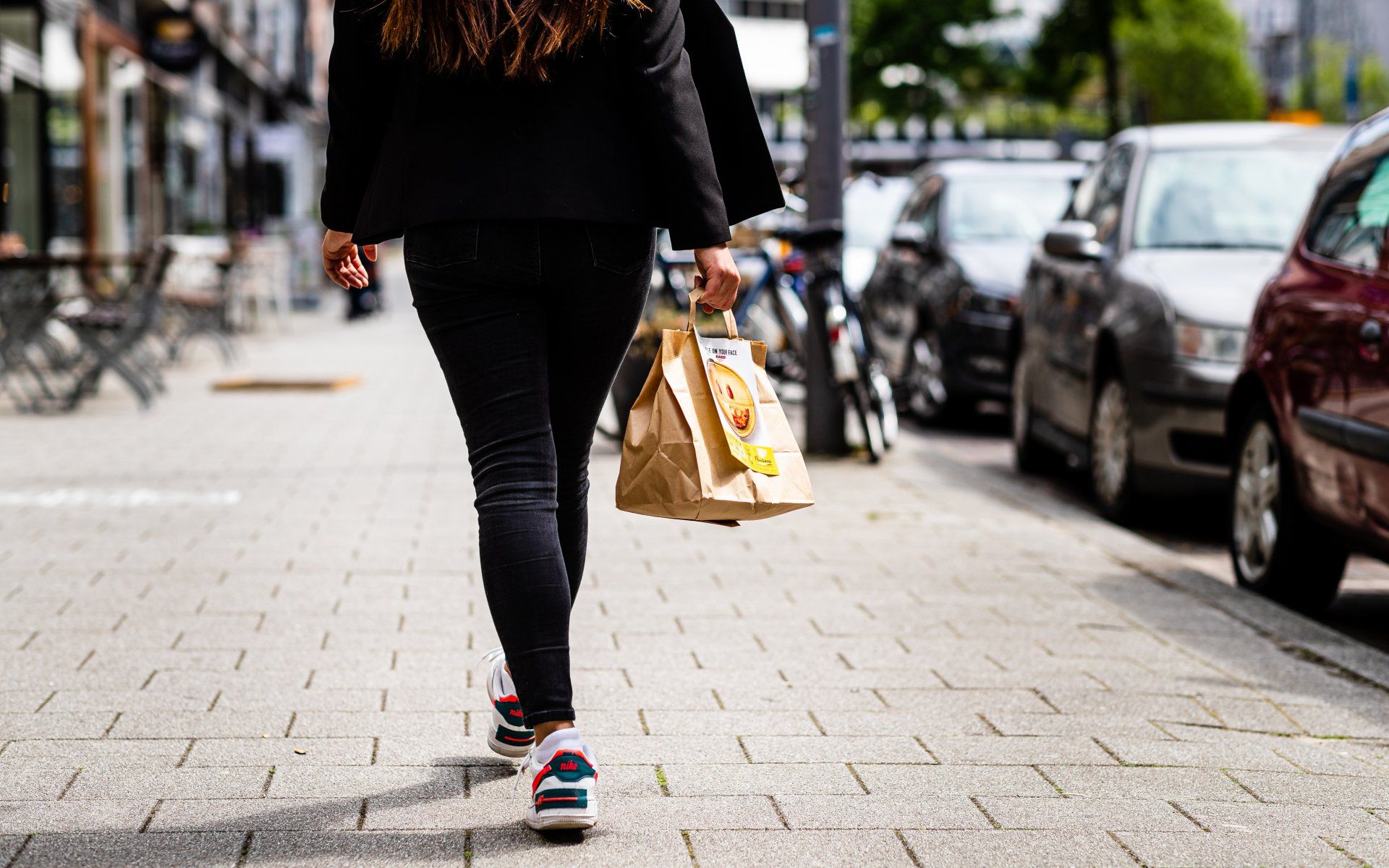 Vrouw loopt over de stoep met een bruine papieren tas. Op de achtergrond staan auto's geparkeerd op straat.