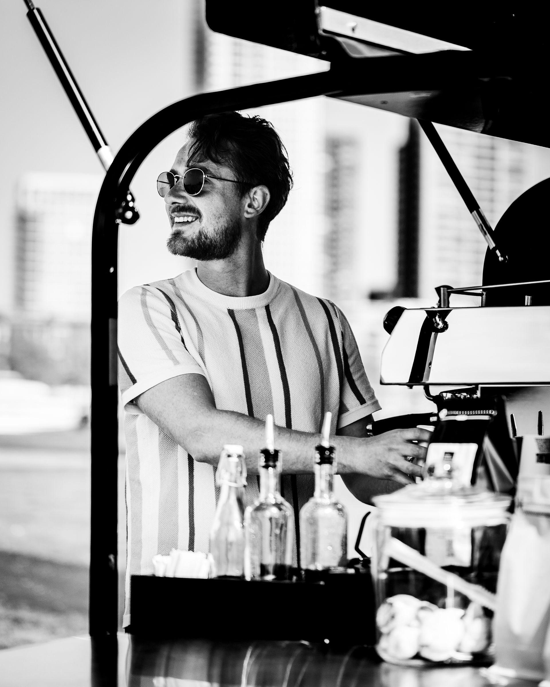Een glimlachende man leunt tegen een koffiekarretje en kijkt opzij. Hij draagt een zonnebril en een gestreept shirt.