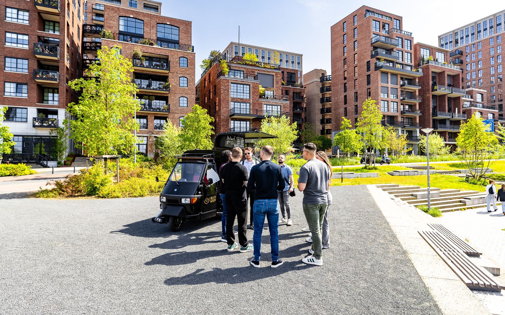Mensen verzamelen zich rond een zwart voertuig op een plein met moderne bakstenen gebouwen en groen op de achtergrond.