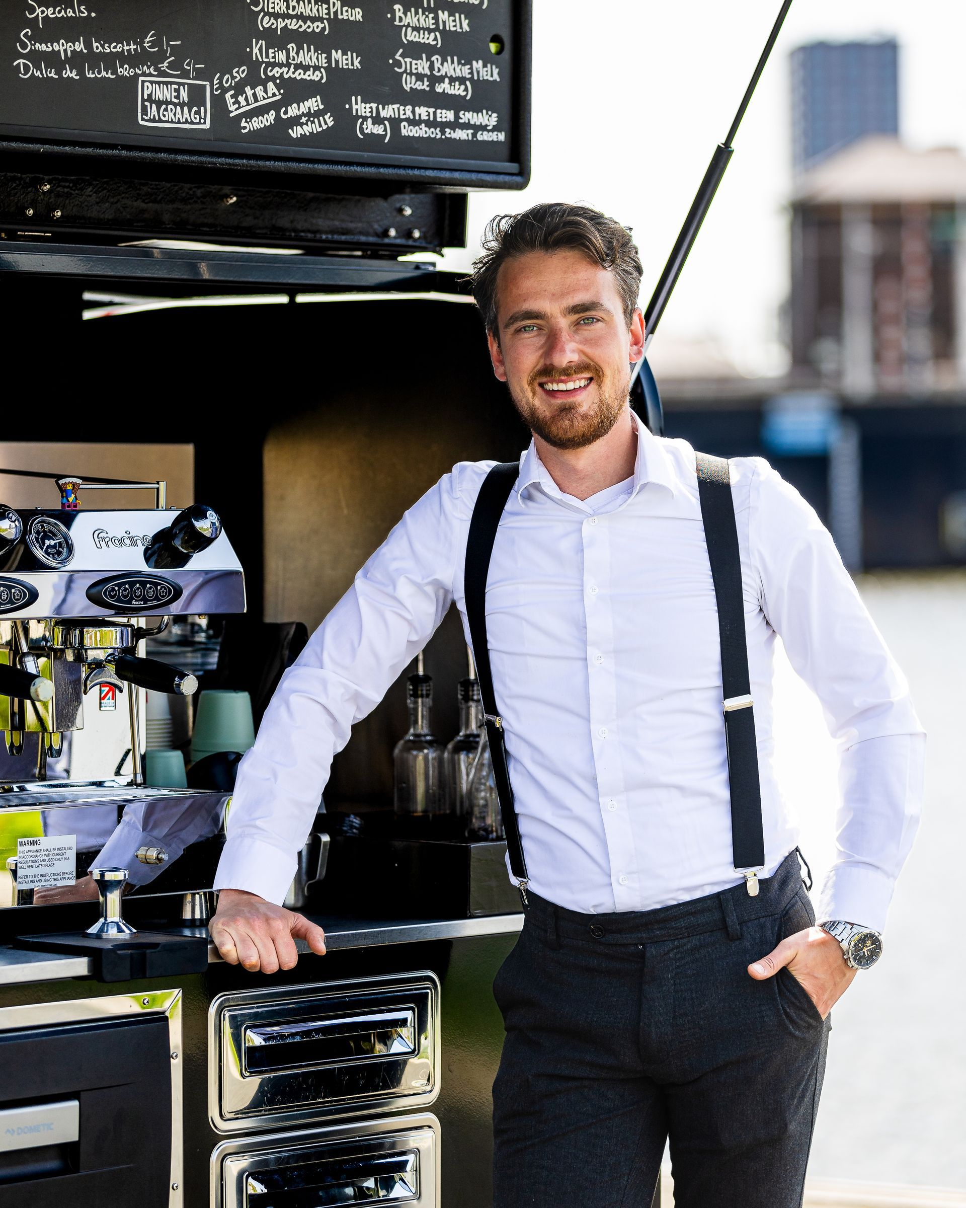 Een man in een wit overhemd en bretels leunt glimlachend tegen een koffiekarretje.