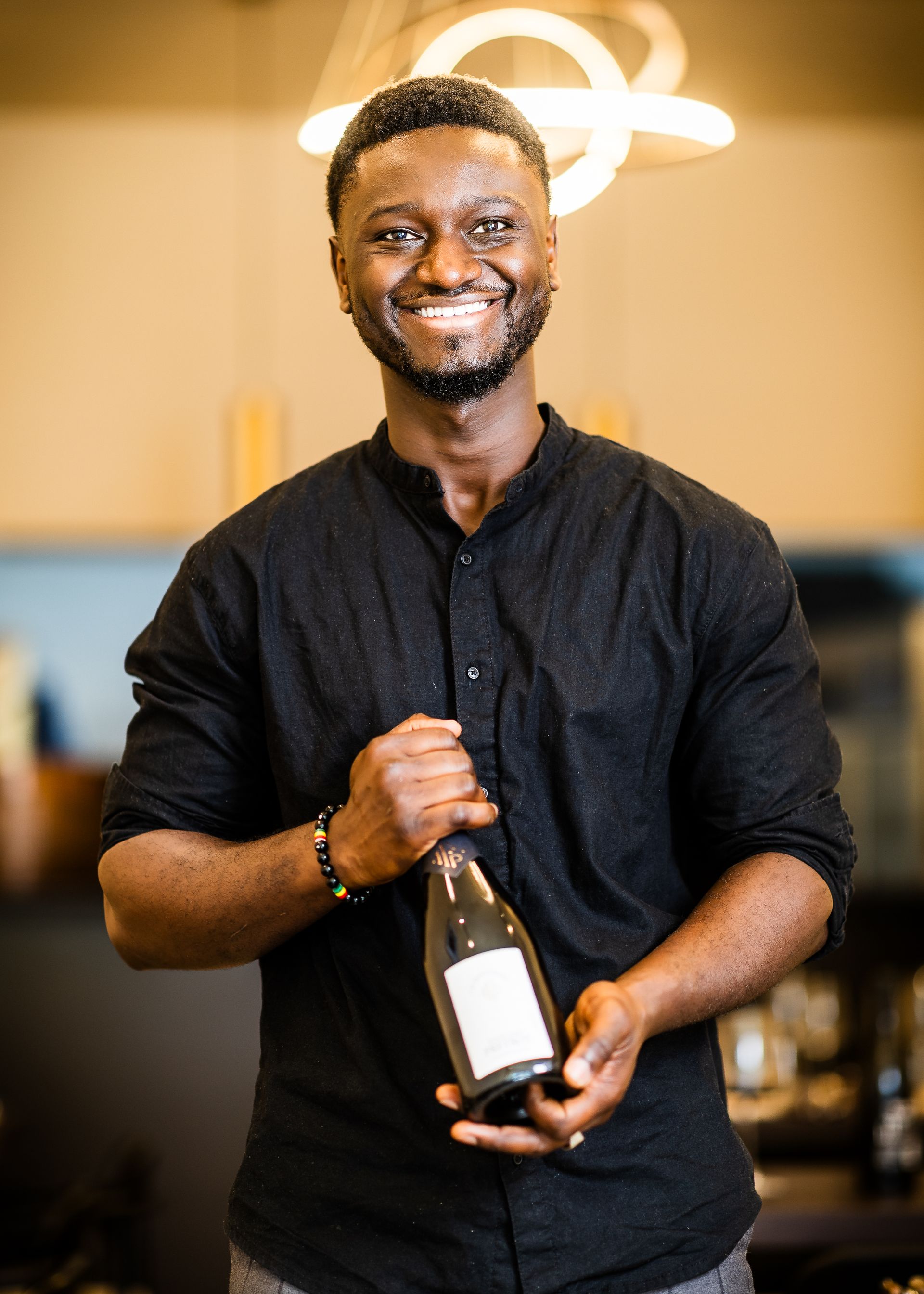 Een man in een zwart shirt glimlacht terwijl hij een fles met een blanco etiket vasthoudt, binnen met een neutrale achtergrond.
