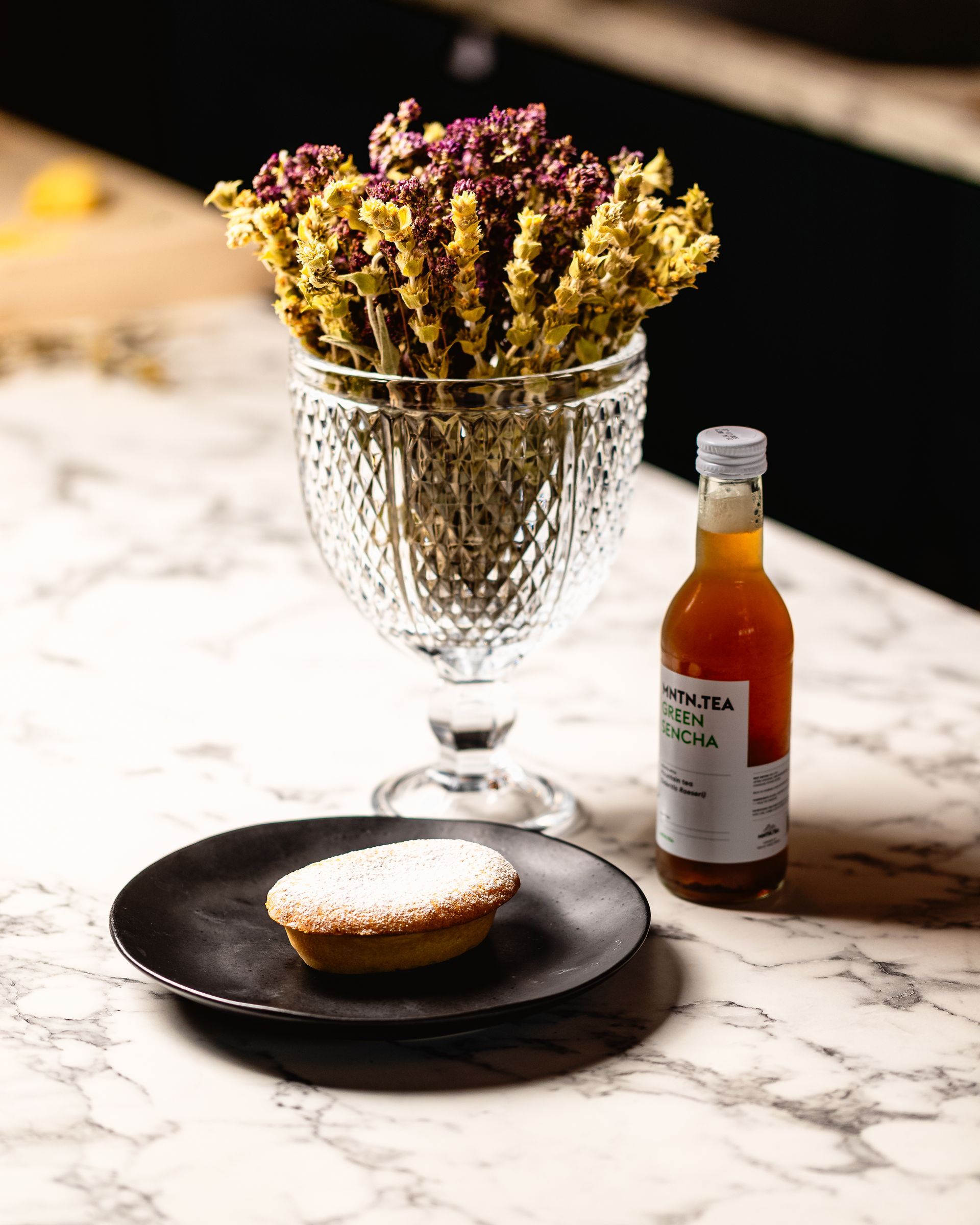 Een taart op een zwart bord, een fles thee en een glazen vaas met gedroogde bloemen op een marmeren blad.