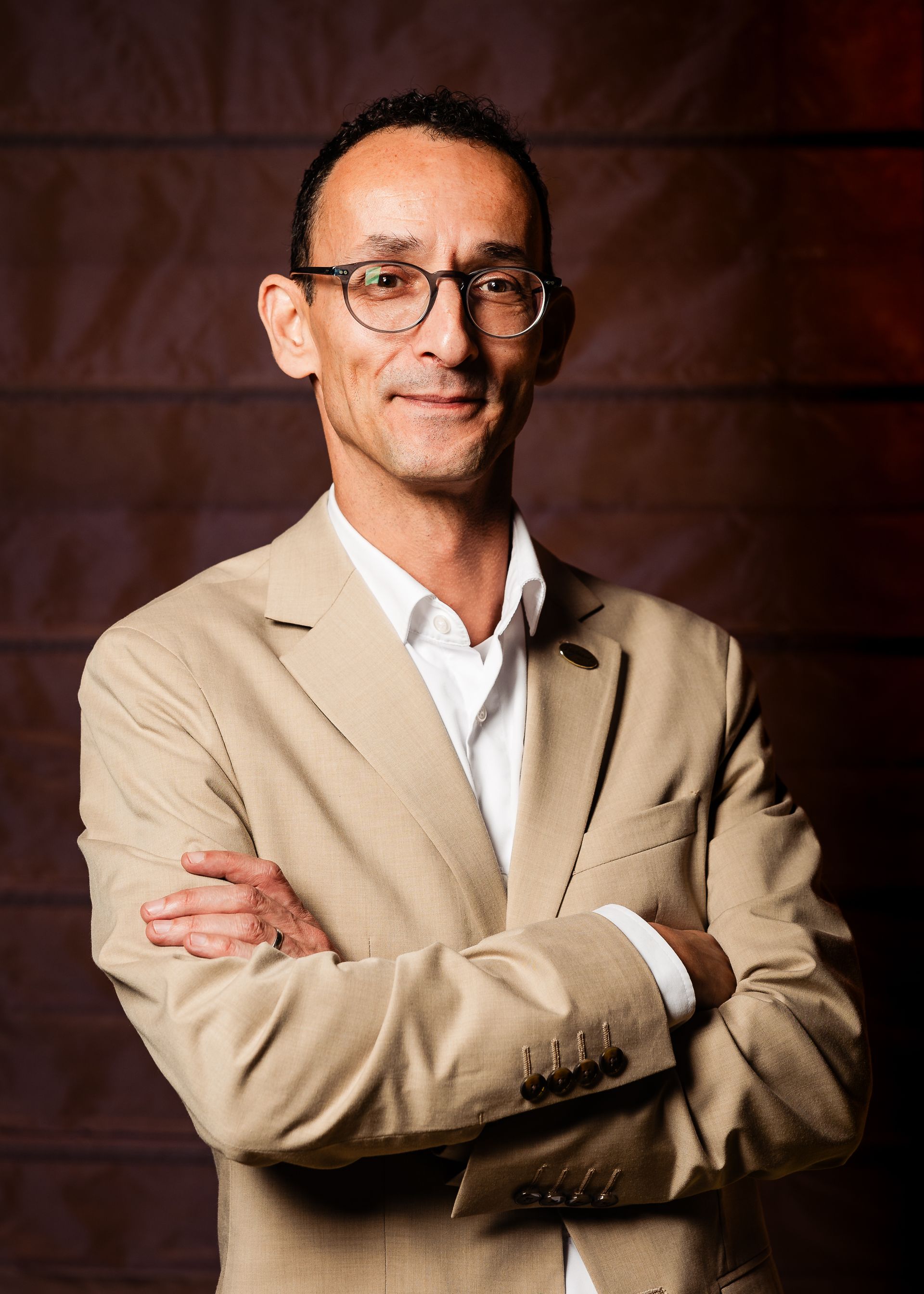 Een man met een bril, een beige blazer en een wit overhemd staat met zijn armen over elkaar voor een bruine muur.