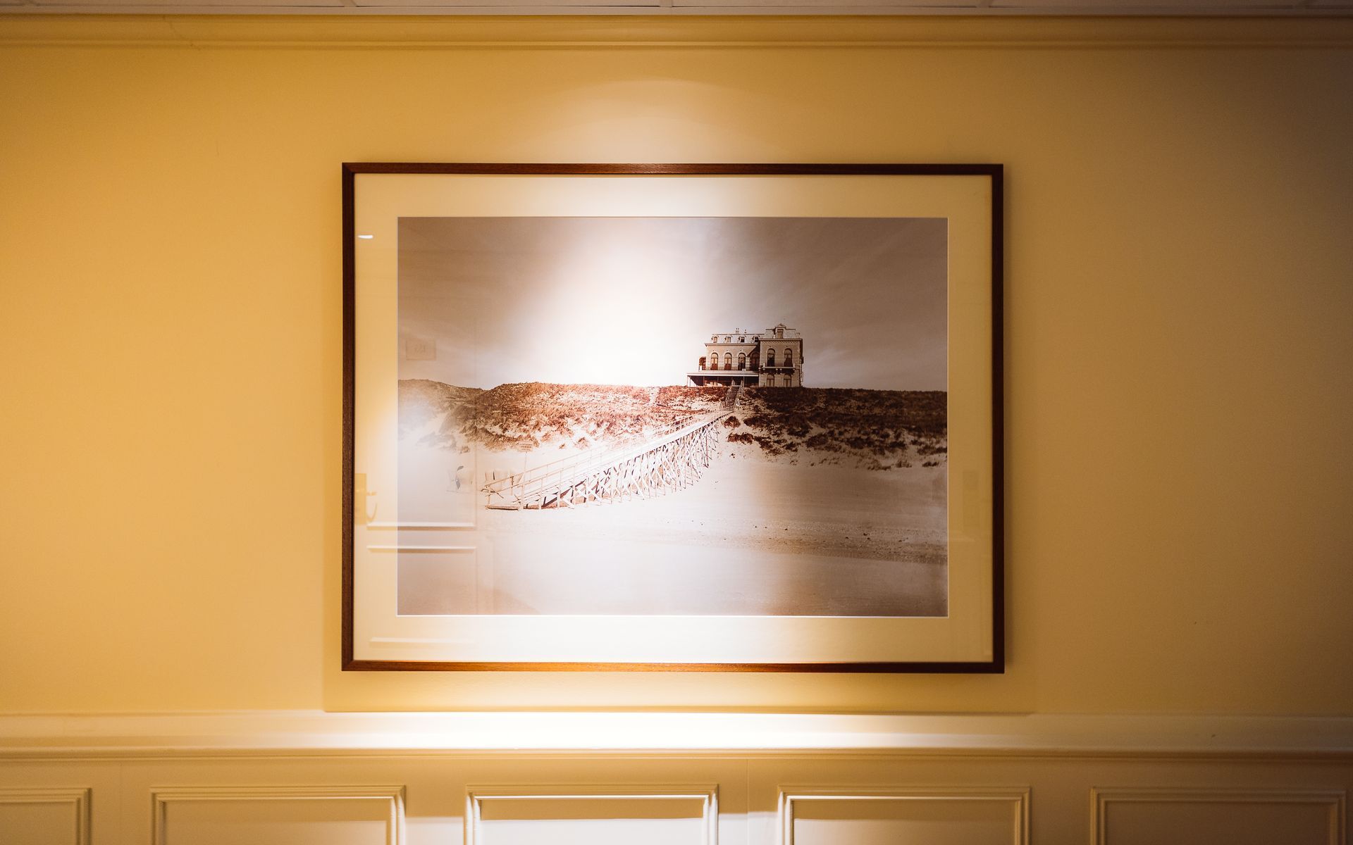 Ingelijste sepia-afdruk van een kustgebouw aan een muur, fel verlicht, met sierlijst aan de onderkant.