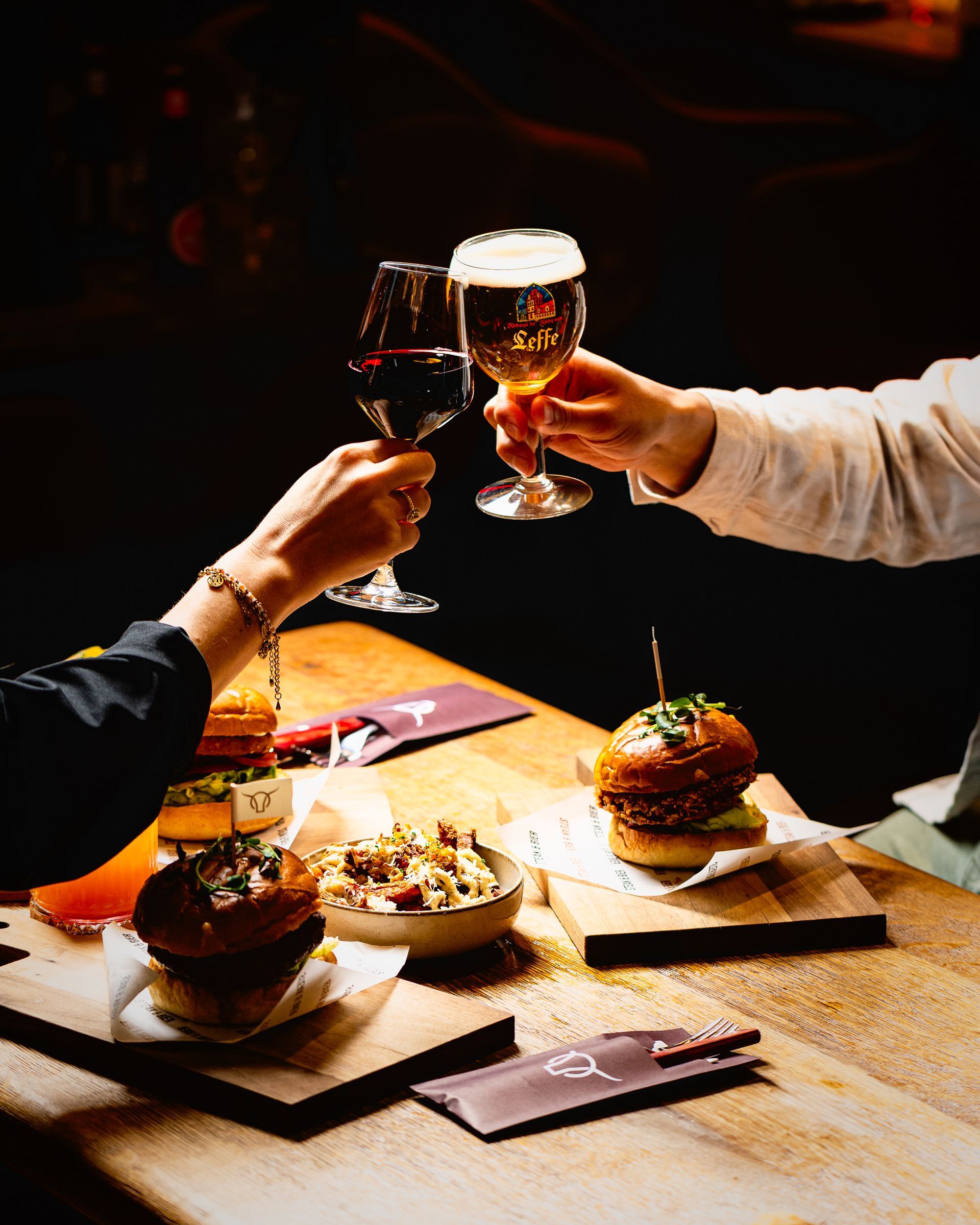 Twee mensen proosten met wijn en bier aan een tafel met hamburgers en bijgerechten.