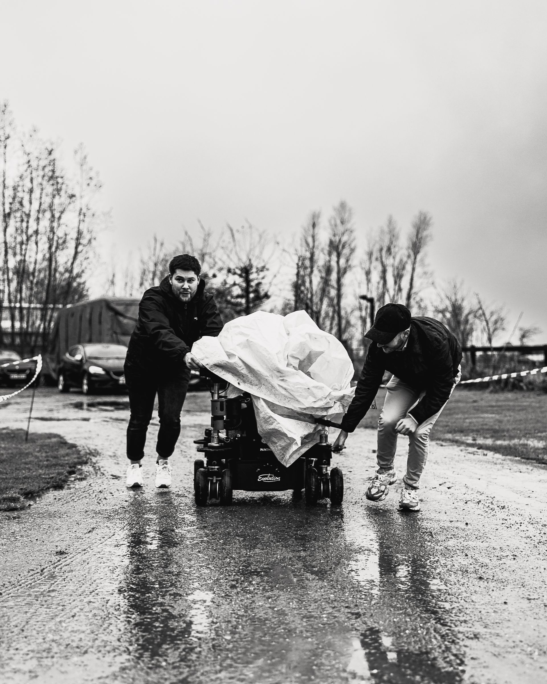 Twee mensen duwen een met plastic bedekte rolstoel over een nat pad; het is een bewolkte dag.
