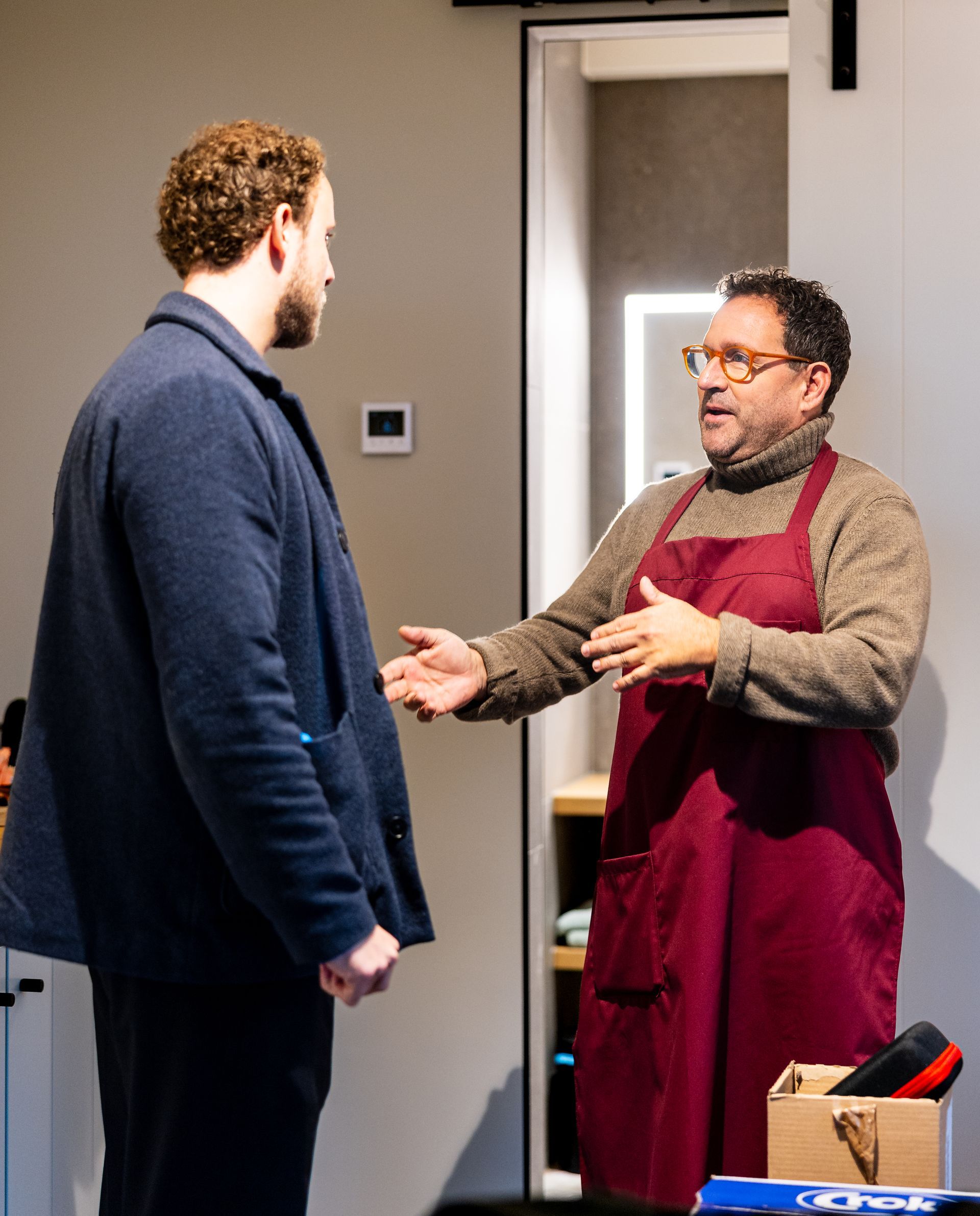 Twee mannen in gesprek in een neutrale kamer. De ene draagt een donkere jas, de andere een schort.