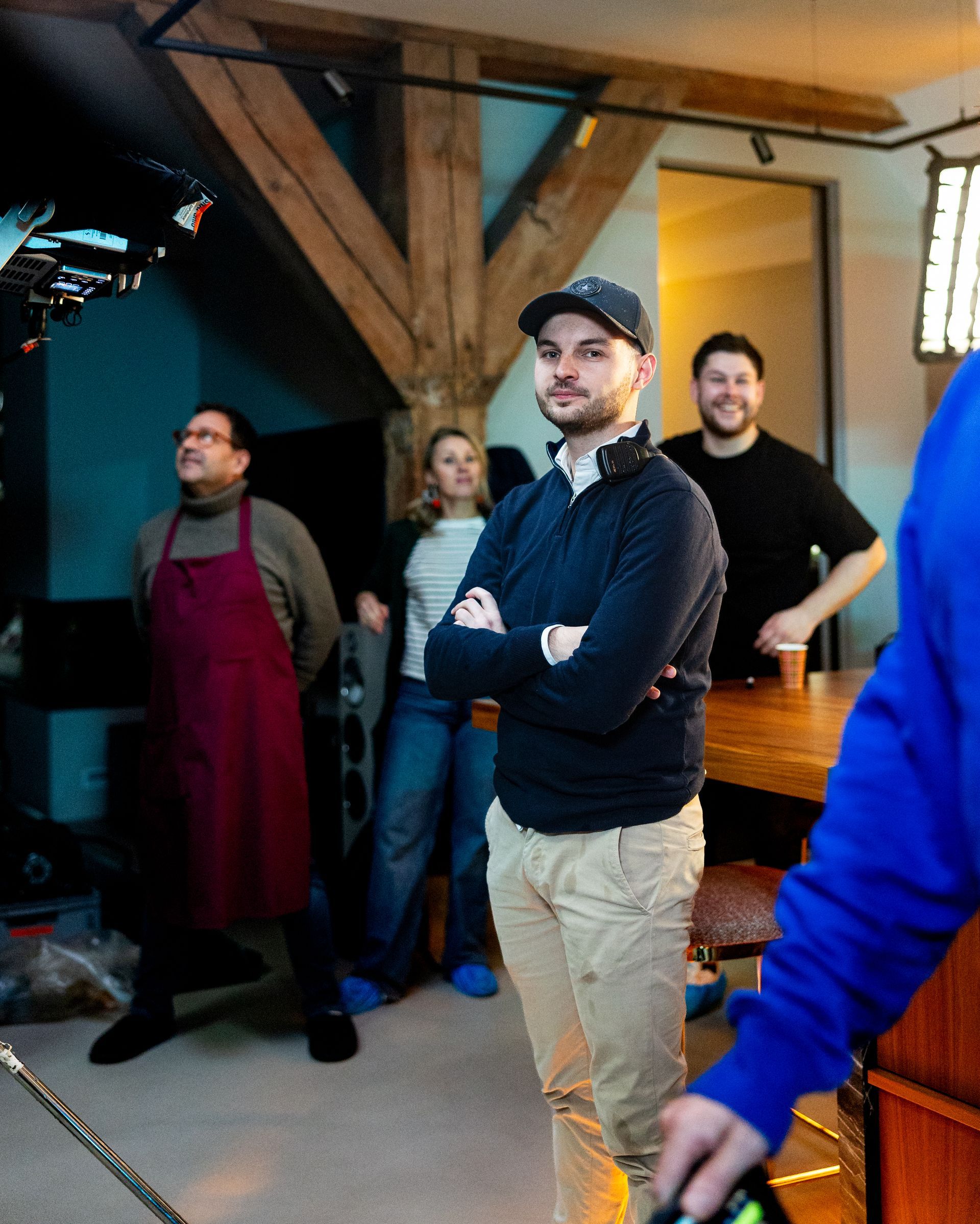 Mensen op een filmset. Een man in het midden met de armen over elkaar, draagt een pet en een donkere trui. Anderen kijken toe, sommigen bij een tafel.