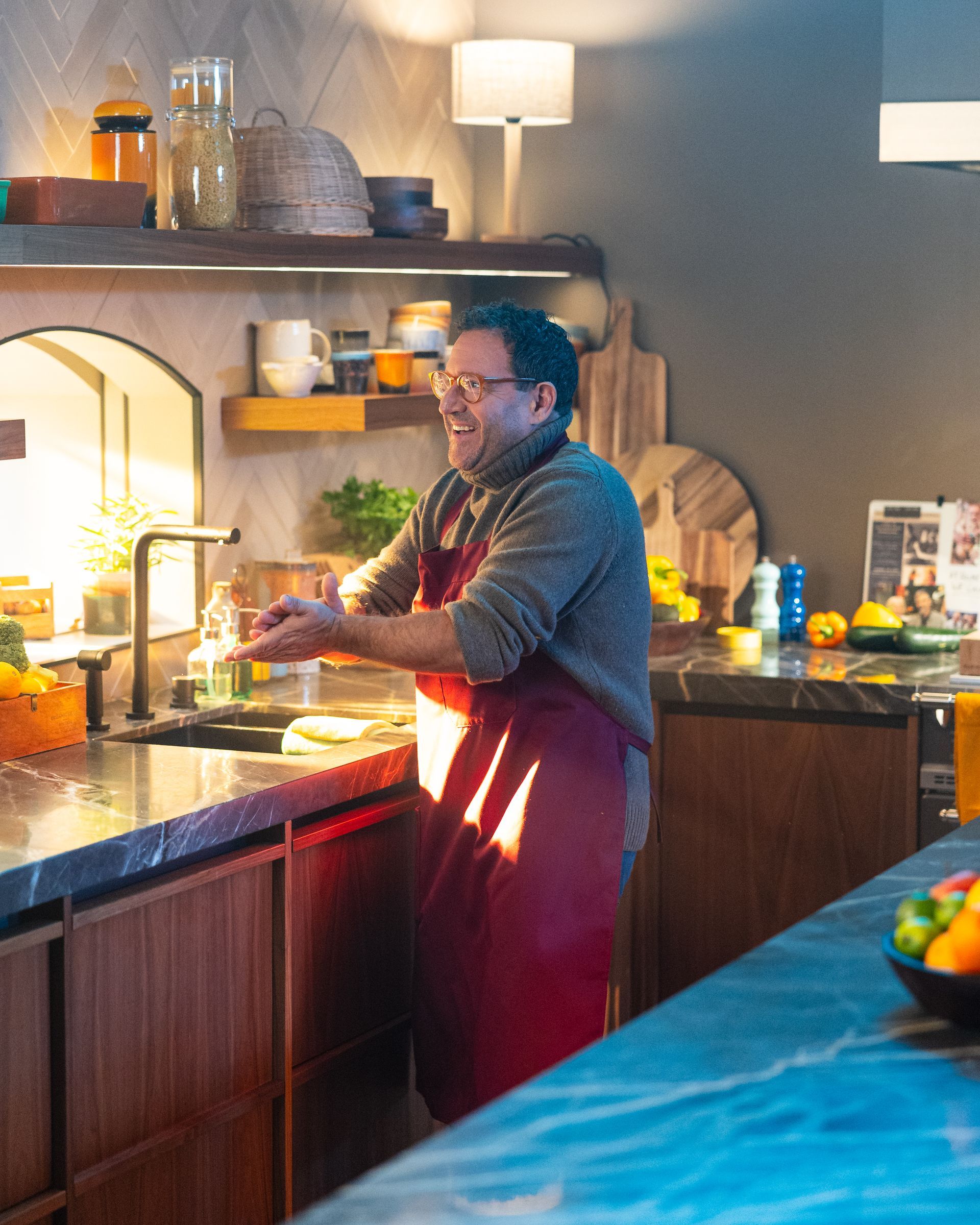 Een man wast zijn handen in de keuken, met een rood schort om. Verlicht aanrecht, sinaasappels en planken.