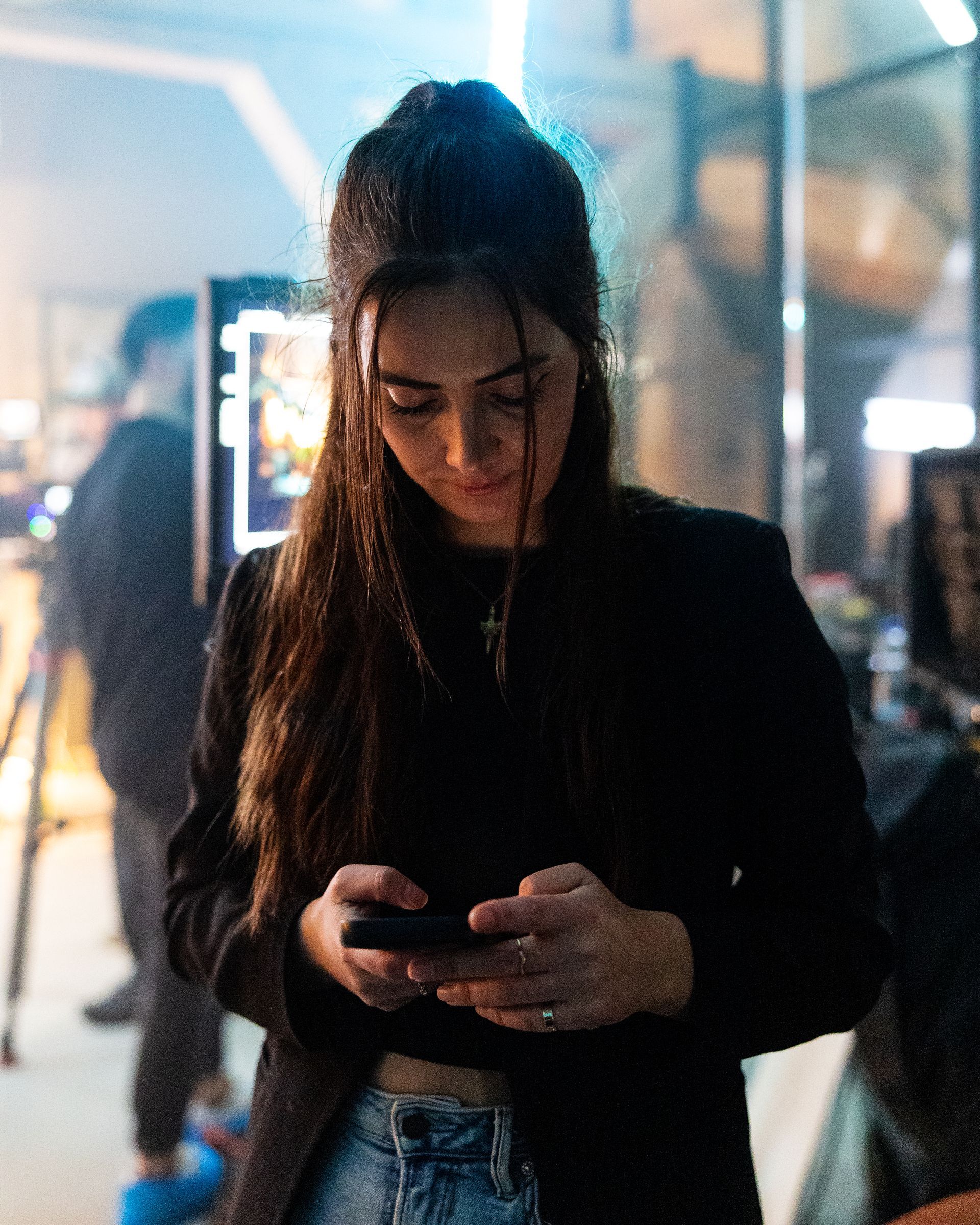 Een vrouw in een zwarte blazer kijkt naar haar telefoon in een ruimte vol technologie, verlicht met blauwe en oranje lampen.