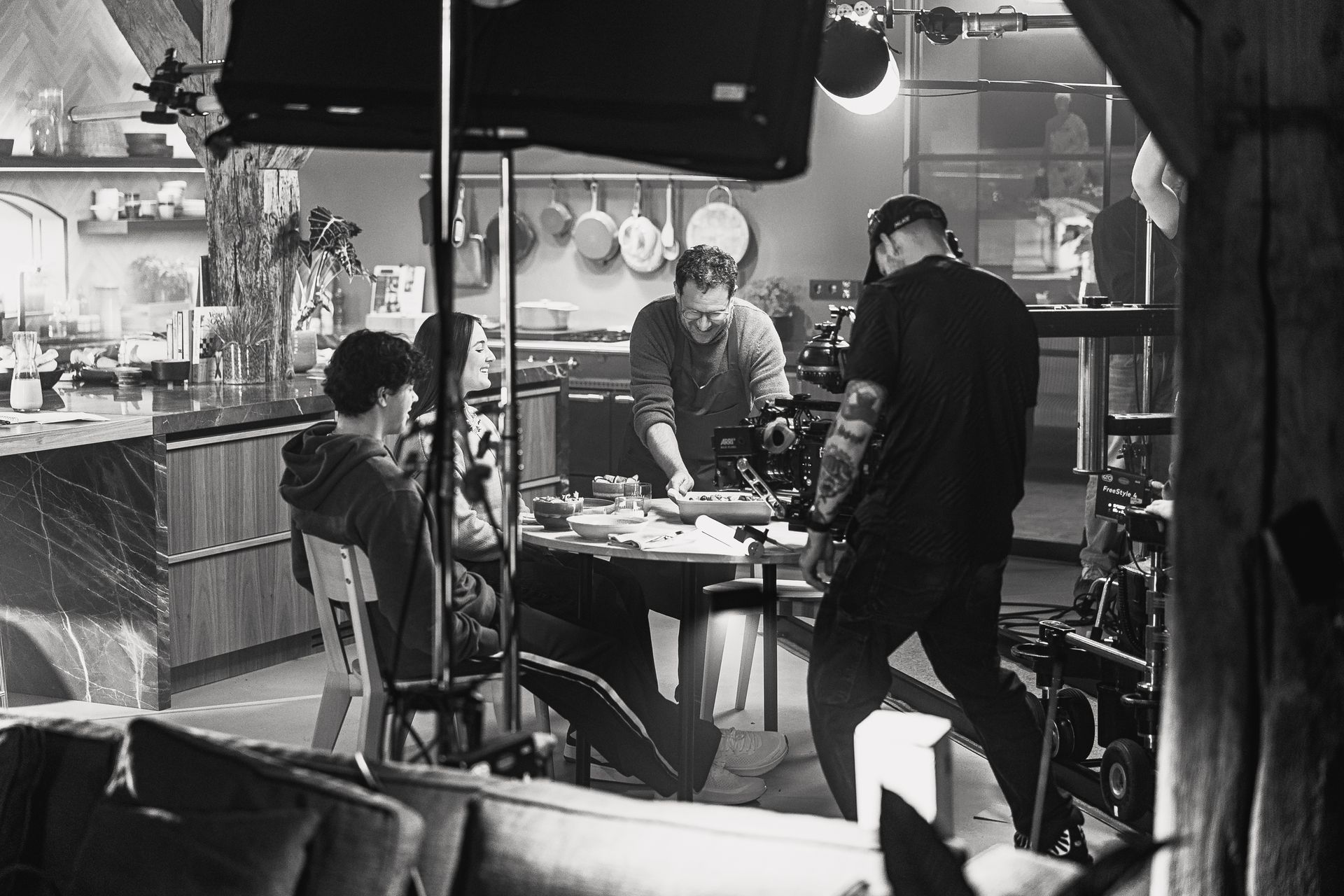 Een filmploeg is aan het filmen in een keuken. Iemand zit, een ander werkt aan een tafel en een cameraman filmt. Zwart-wit.