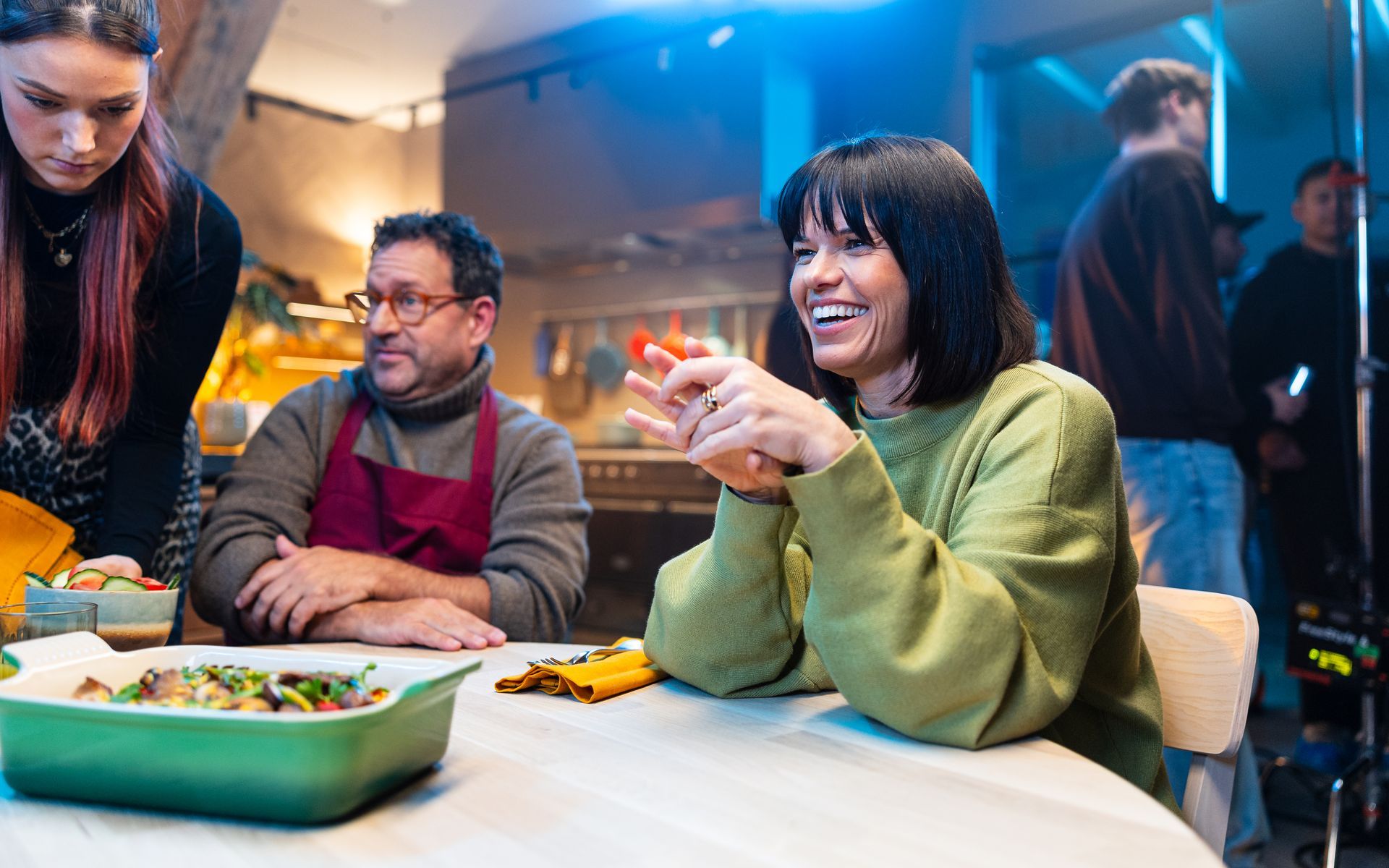 Een vrouw glimlacht aan een tafel met eten. Een man met een schort kijkt toe. Op de achtergrond is een filmploeg te zien, in een keukenomgeving.