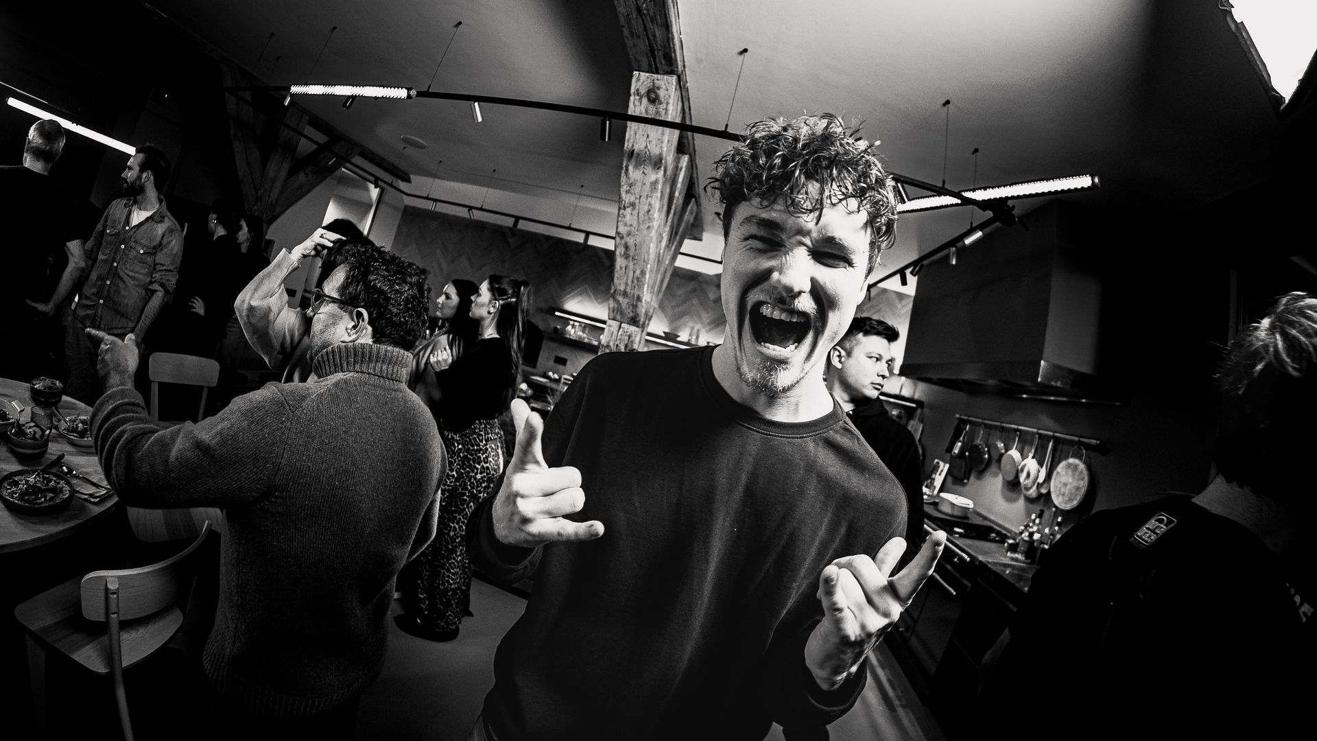 Een opgewonden man met krullend haar steekt zijn duim omhoog en schreeuwt op een feestje. Zwart-witfoto.