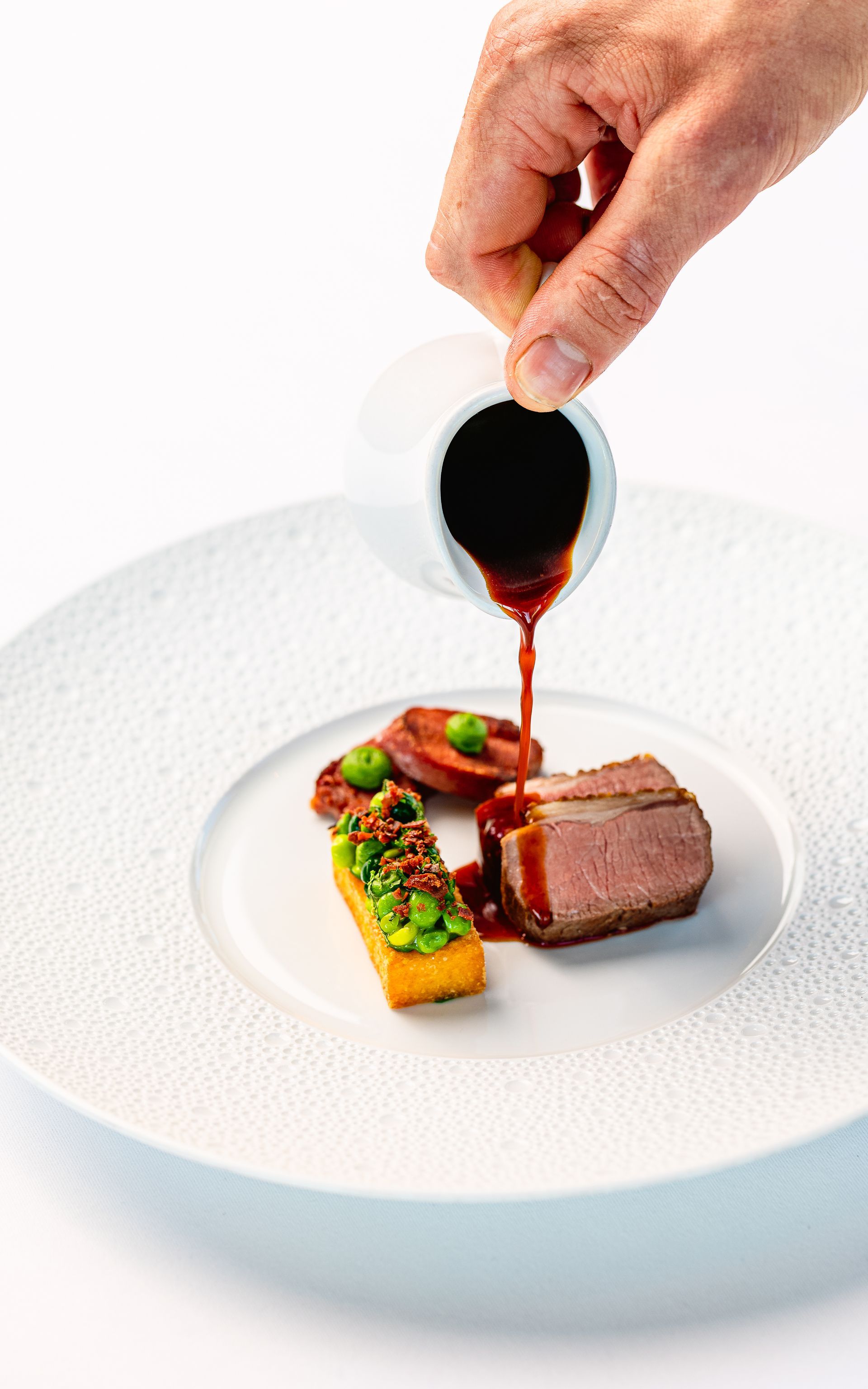 Een hand giet saus over een bord met gastronomische gerechten. Inclusief biefstuk, groenten en saus.