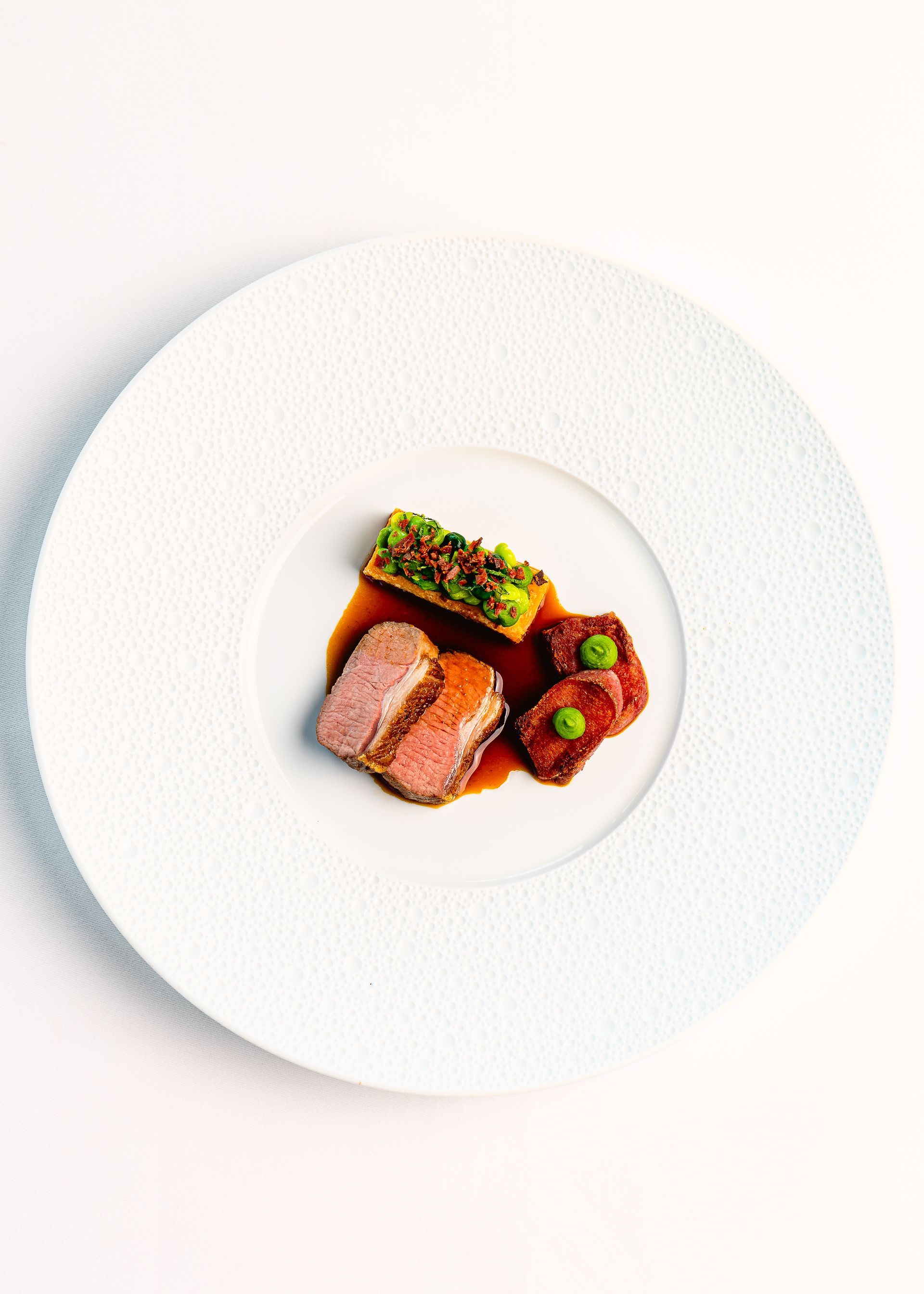 Een gastronomisch gerecht met gesneden vlees, een bijgerecht van groenten en saus op een wit, gestructureerd bord.
