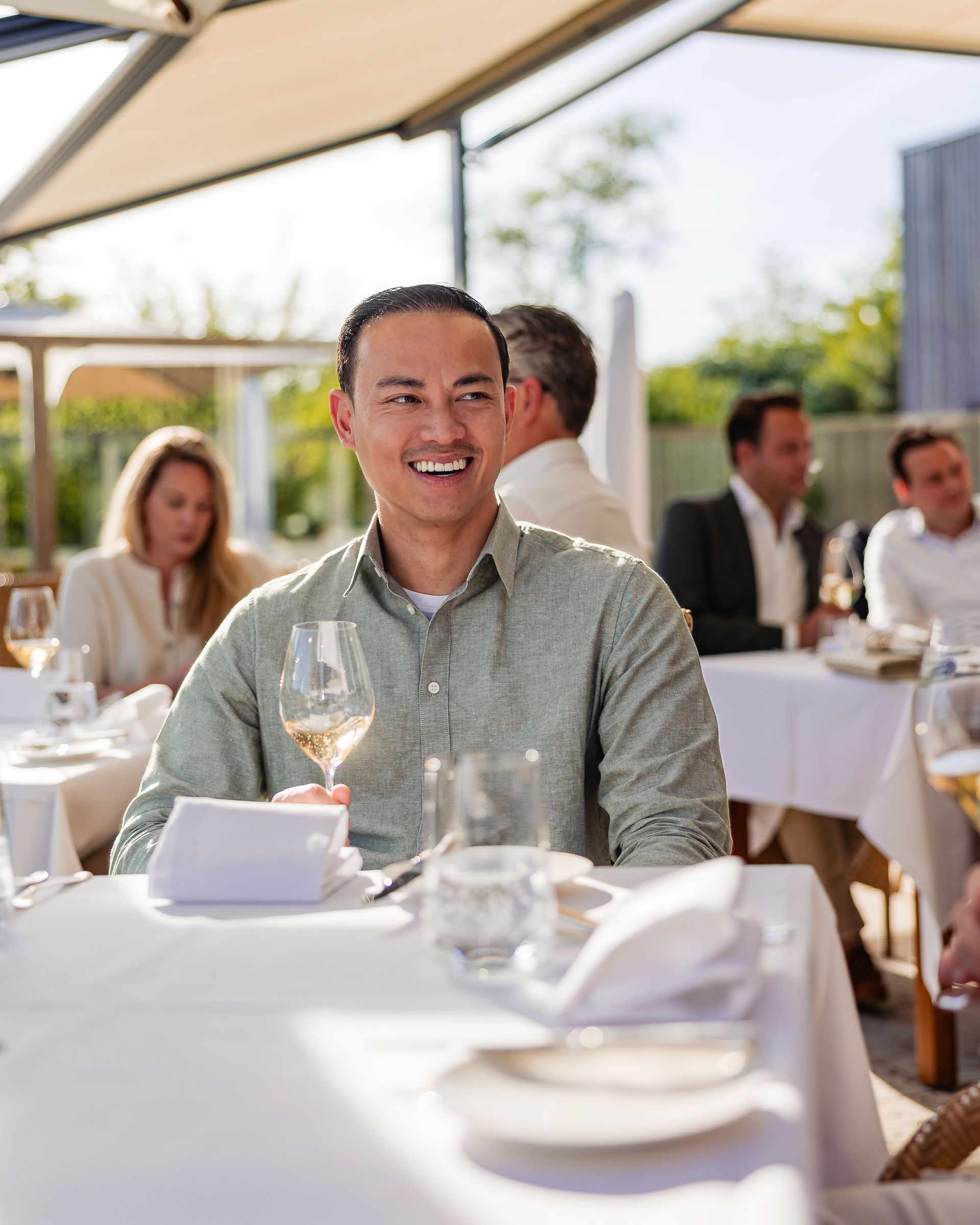 Een glimlachende man met een wijnglas in zijn hand zit met anderen aan een tafeltje in een restaurant op het terras.