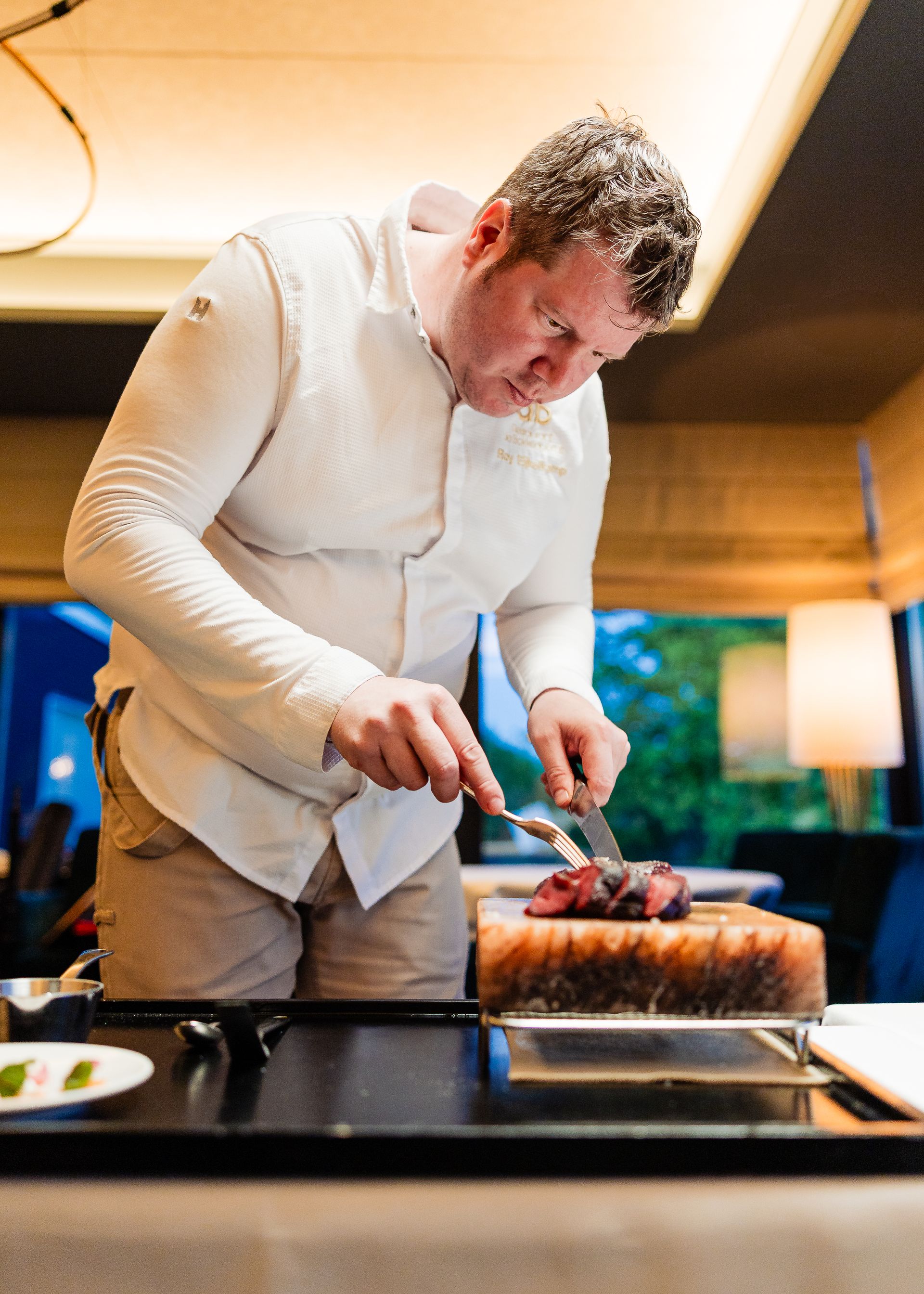 Een chef-kok in wit uniform snijdt voedsel op een houten snijplank. Binnen, met een geconcentreerde uitdrukking op zijn gezicht.