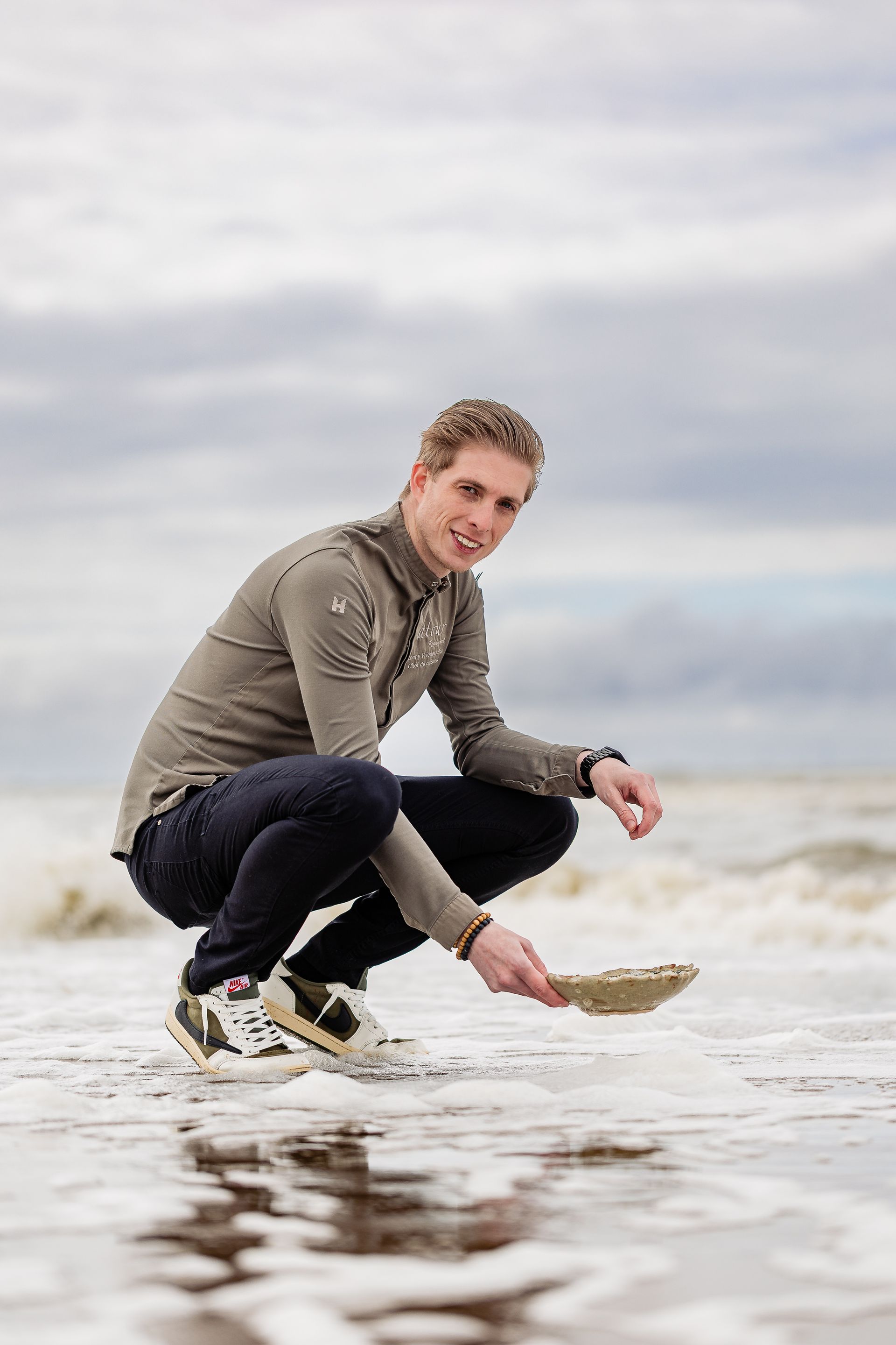 Een man in een olijfgroen shirt en een donkere broek hurkt aan de waterkant, houdt een schelp vast en glimlacht, met de oceaan op de achtergrond.
