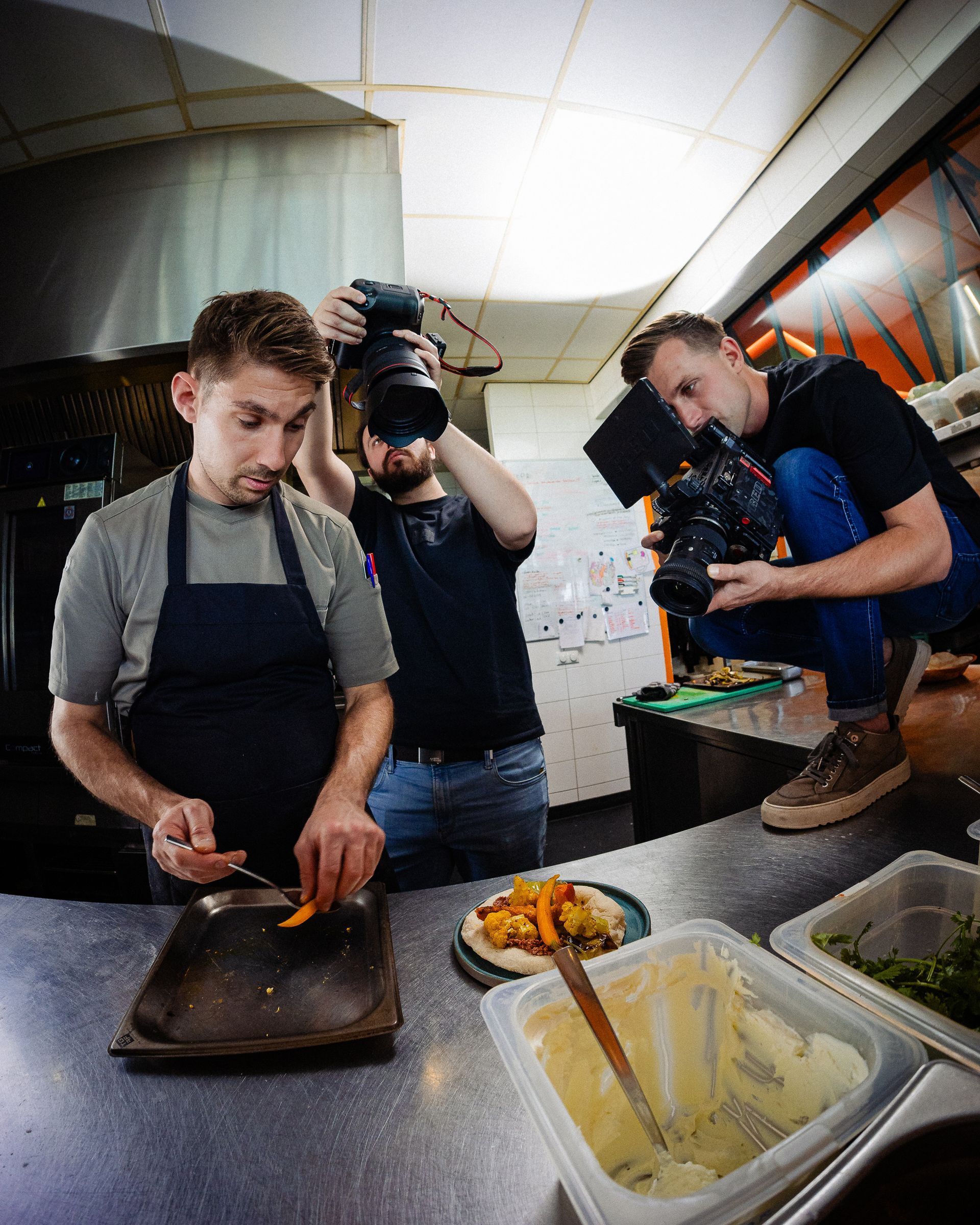 Een chef-kok bereidt eten terwijl twee mensen filmen in een professionele keuken.