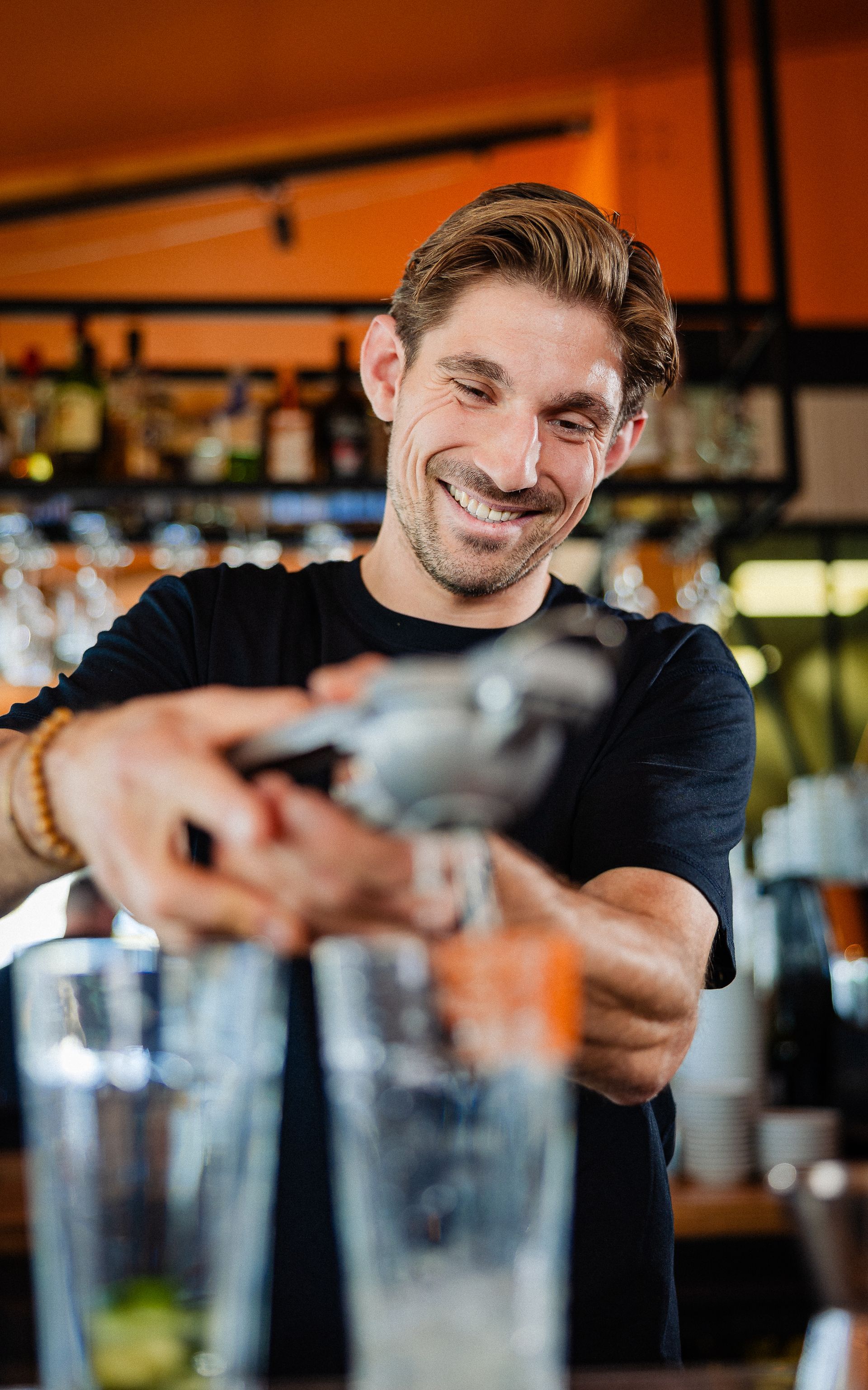 Een barman perst met een glimlach limoen in een shaker. De bar is gedekt met flessen en glazen in het zicht.