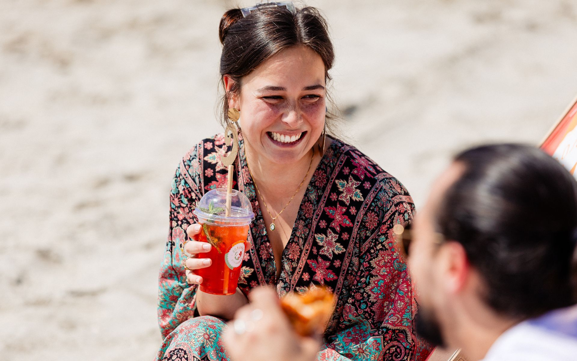 Een vrouw glimlacht, houdt een drankje vast en praat met iemand op het strand.