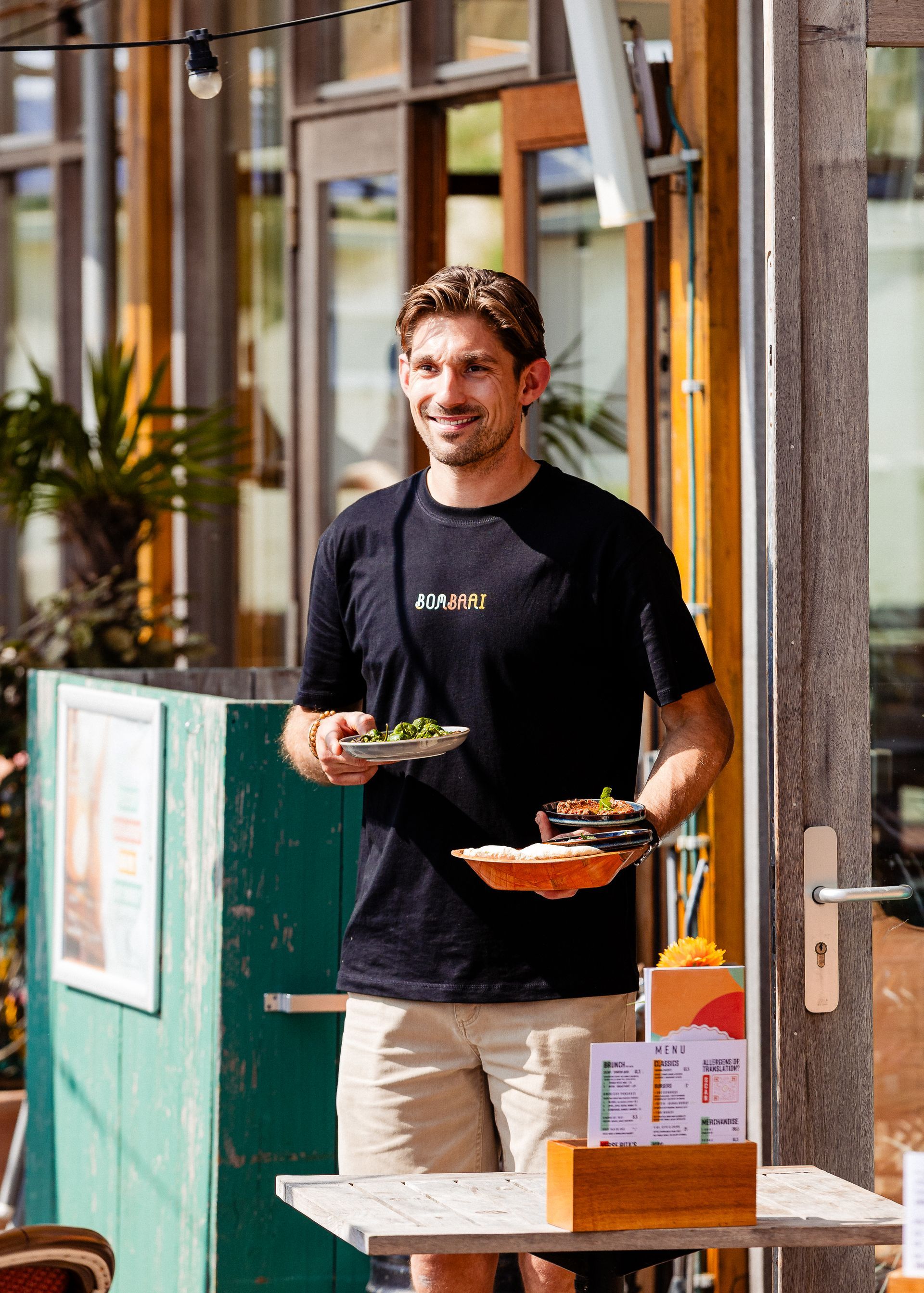 Een man houdt borden met eten vast op een terras bij een restaurant, gekleed in een zwart shirt en een beige korte broek.