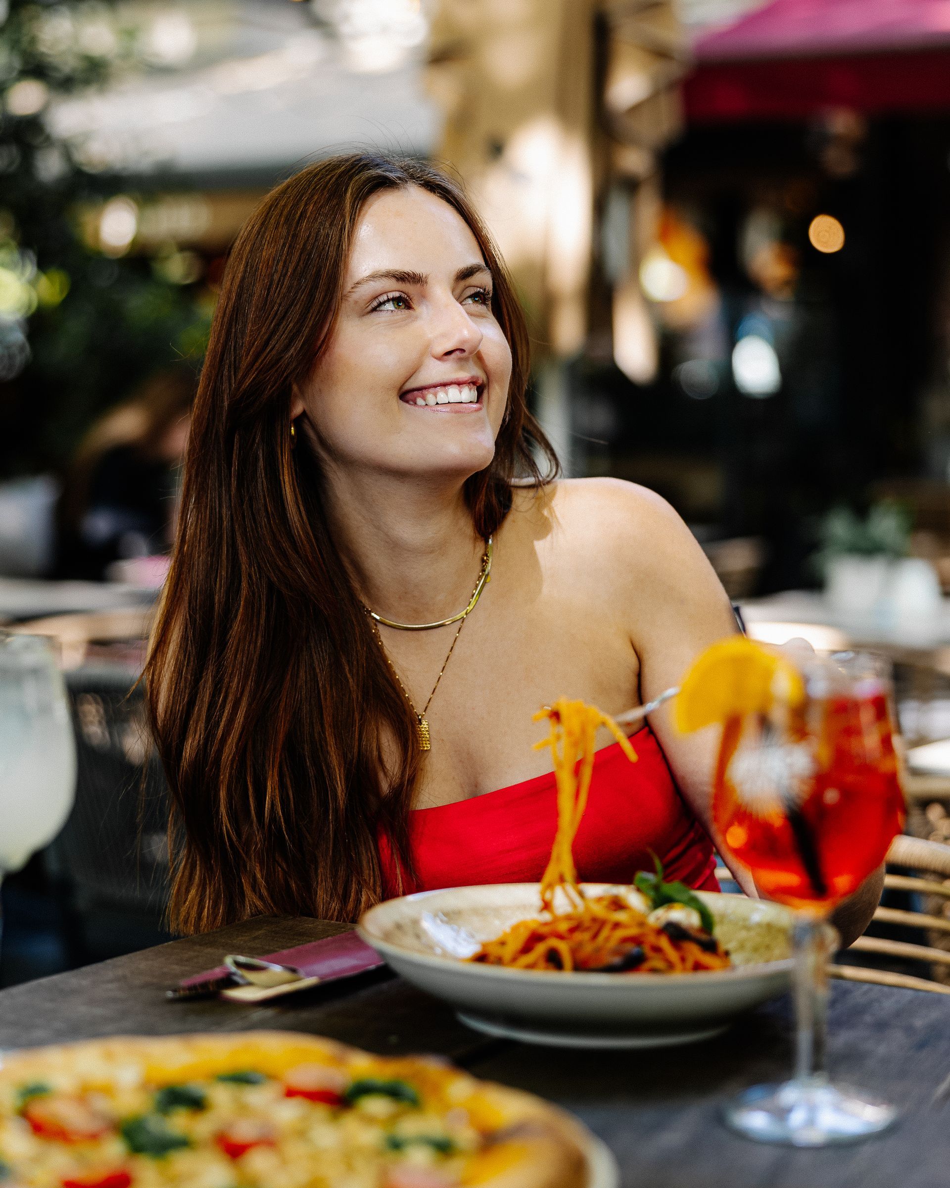 Een lachende vrouw zit aan een tafeltje op het terras van een restaurant. Pasta, een drankje en pizza zijn zichtbaar.