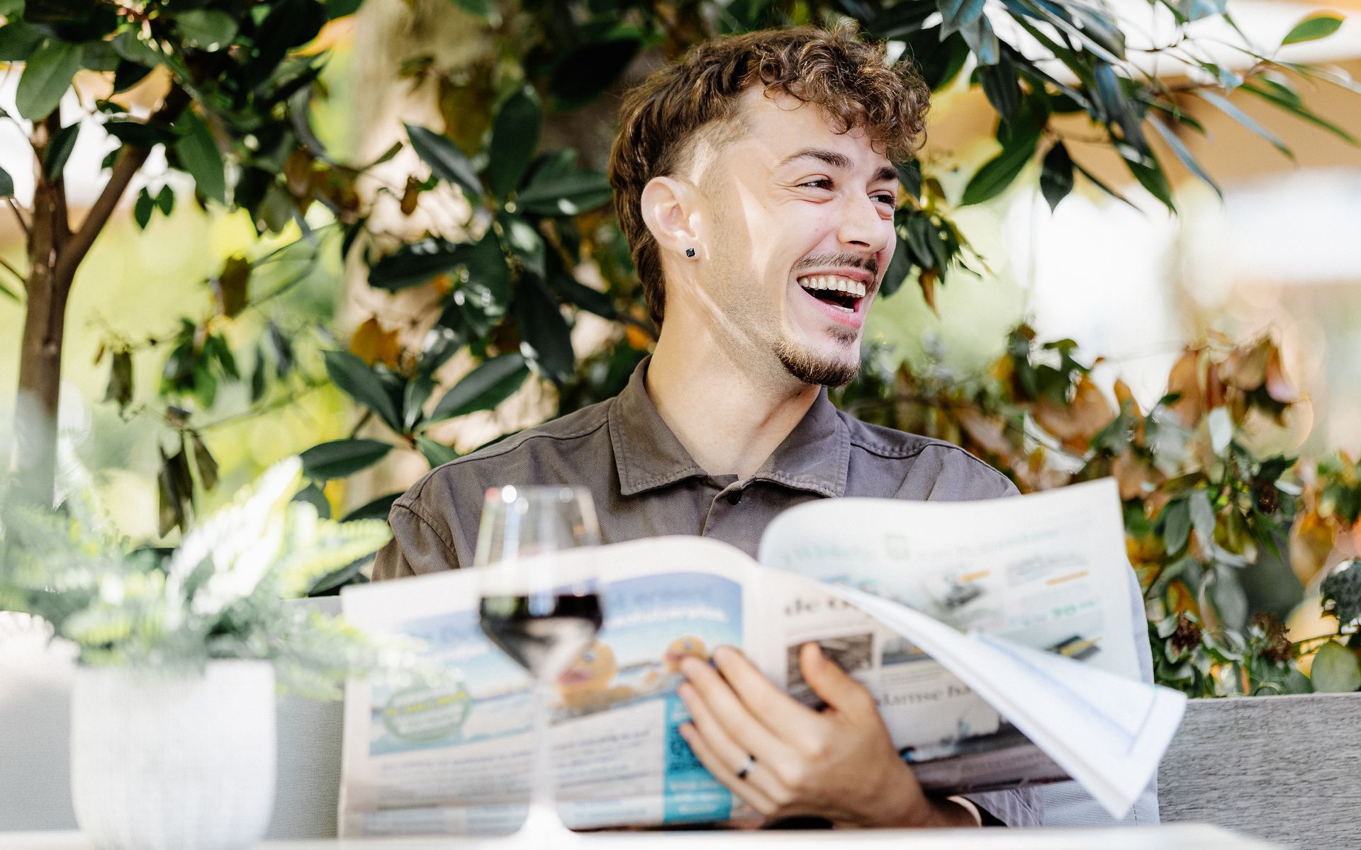 Een lachende man zit aan een buitentafel de krant te lezen, met een glas wijn en planten.