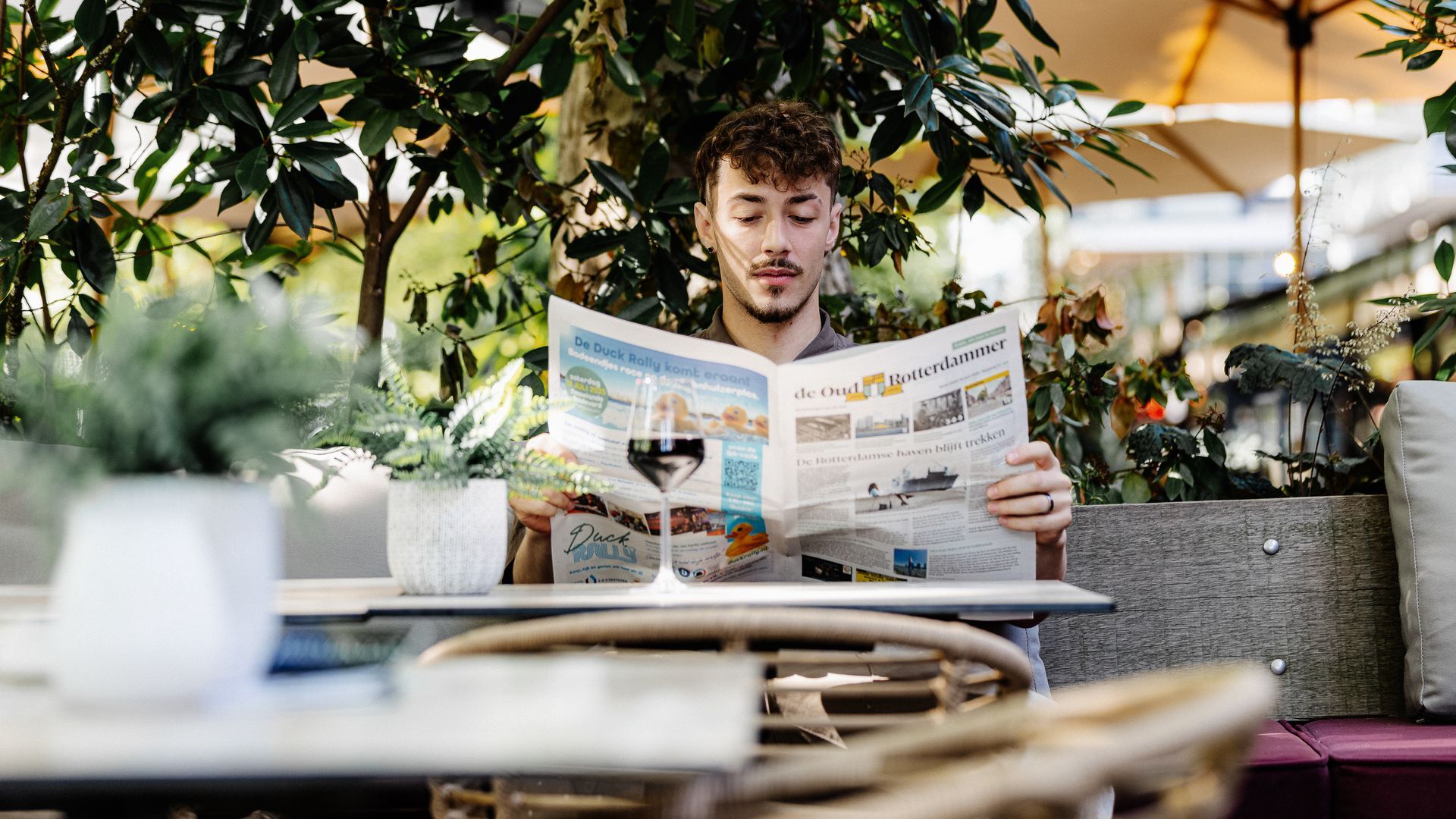 Een man leest buiten aan een tafel de krant met een glas wijn; boven hem is groen te zien.