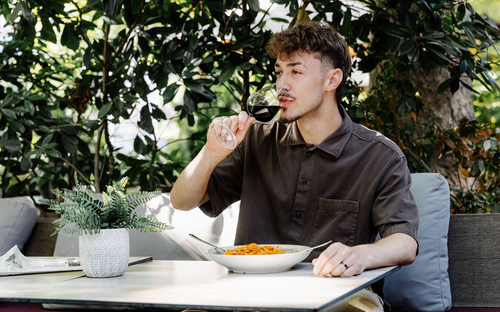 Persoon aan een tafel buiten aan het eten, met een vork in de hand. Witte kom pasta ervoor. Groene bladeren op de achtergrond.