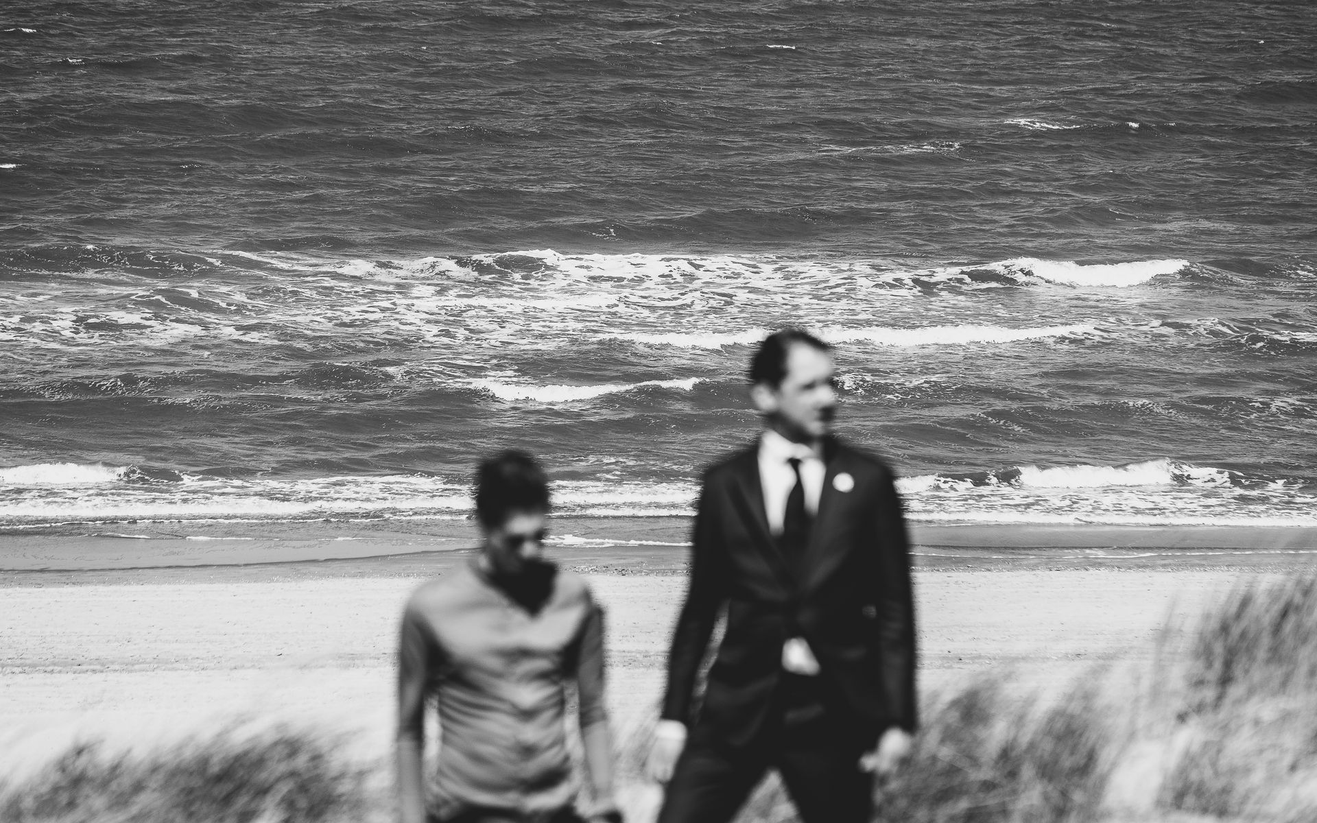 Twee mensen op een strand met de oceaan op de achtergrond. De een in een pak, de ander in een overhemd.