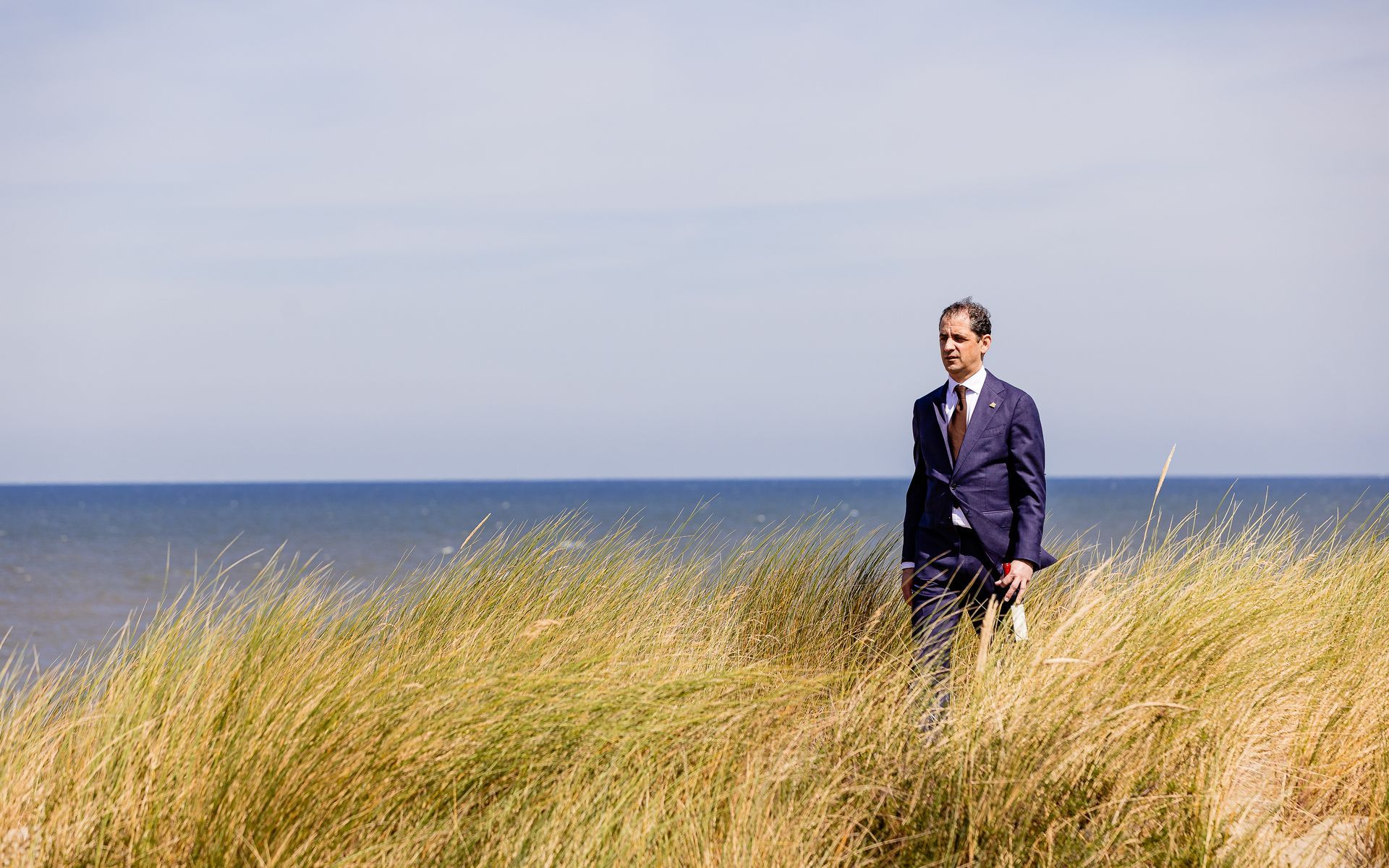 Een man in een pak staat tussen hoog, geel gras met de oceaan op de achtergrond.