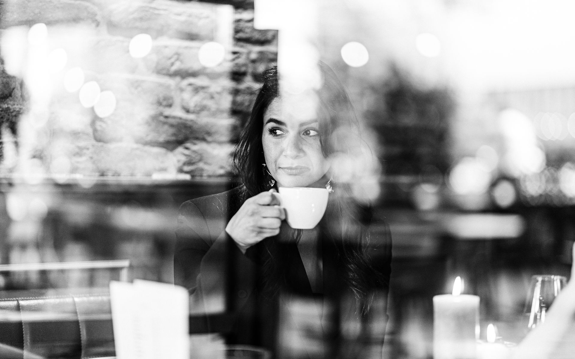 Vrouw in een café, drinkend uit een witte kop. Ze kijkt uit het raam, met op de achtergrond kaarslicht.