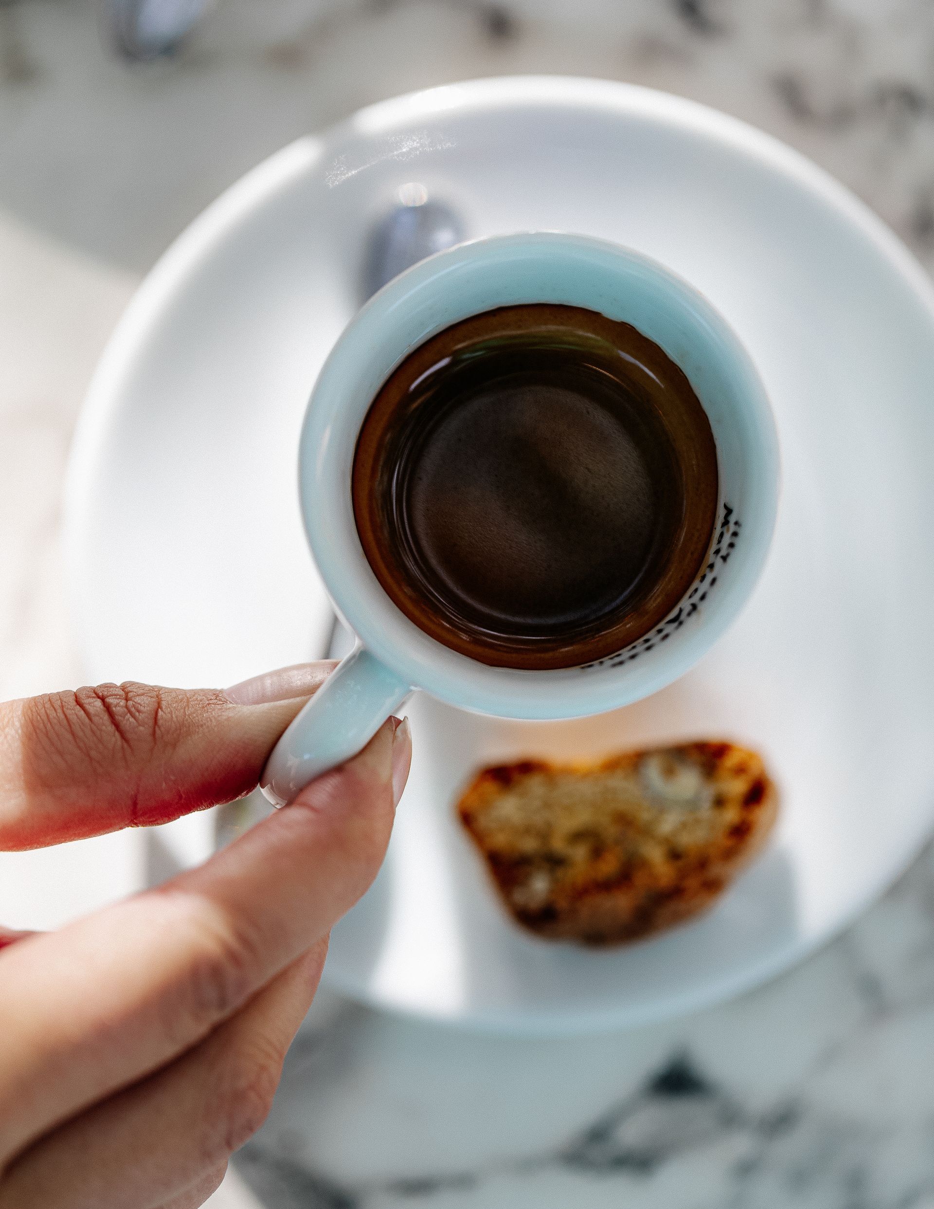 Een hand die een klein kopje espresso vasthoudt op een wit schoteltje met een koekje.
