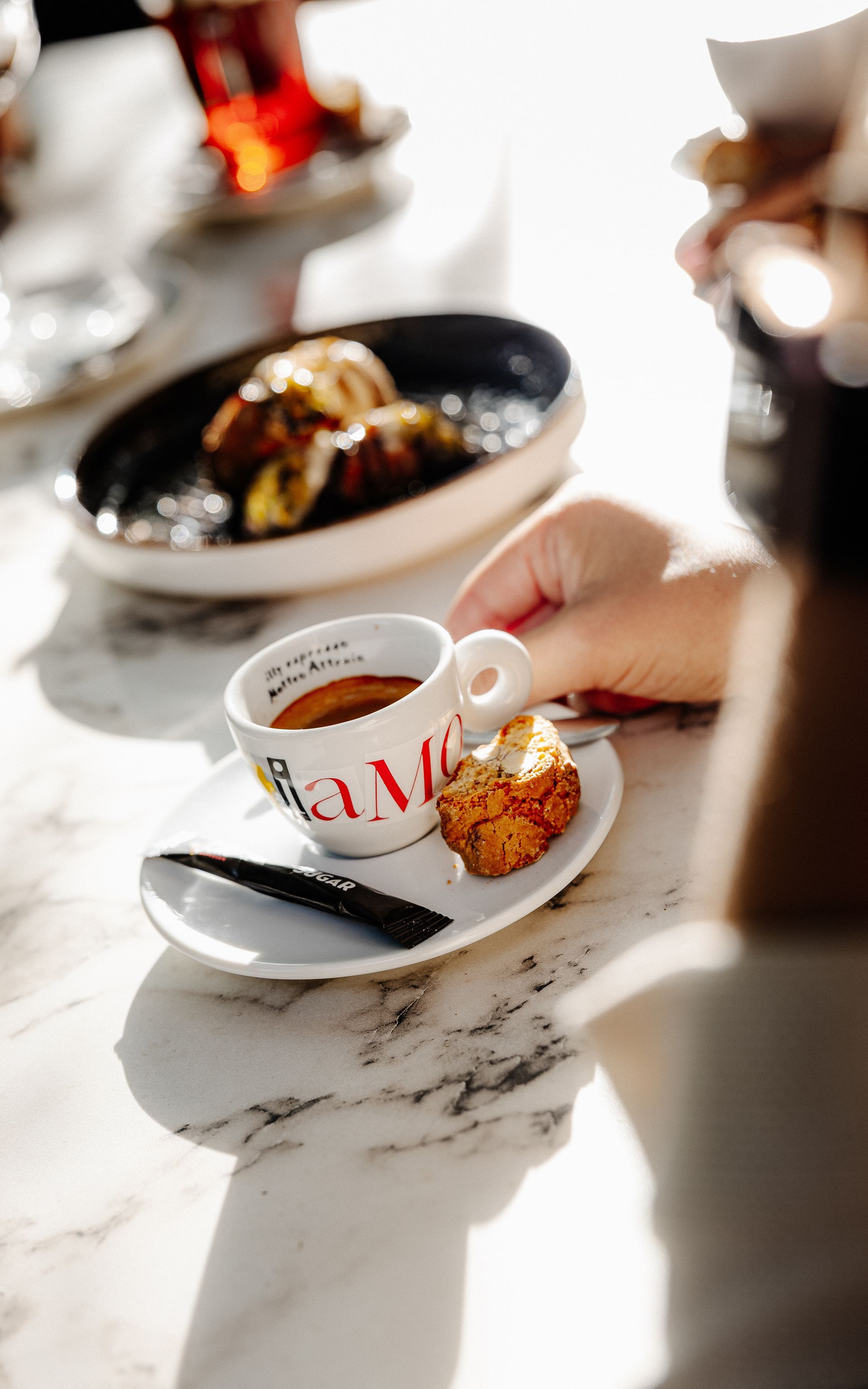 Een persoon houdt een kop koffie en een koekje vast op een wit schoteltje. Op de achtergrond is een zwart bord met gebak te zien.