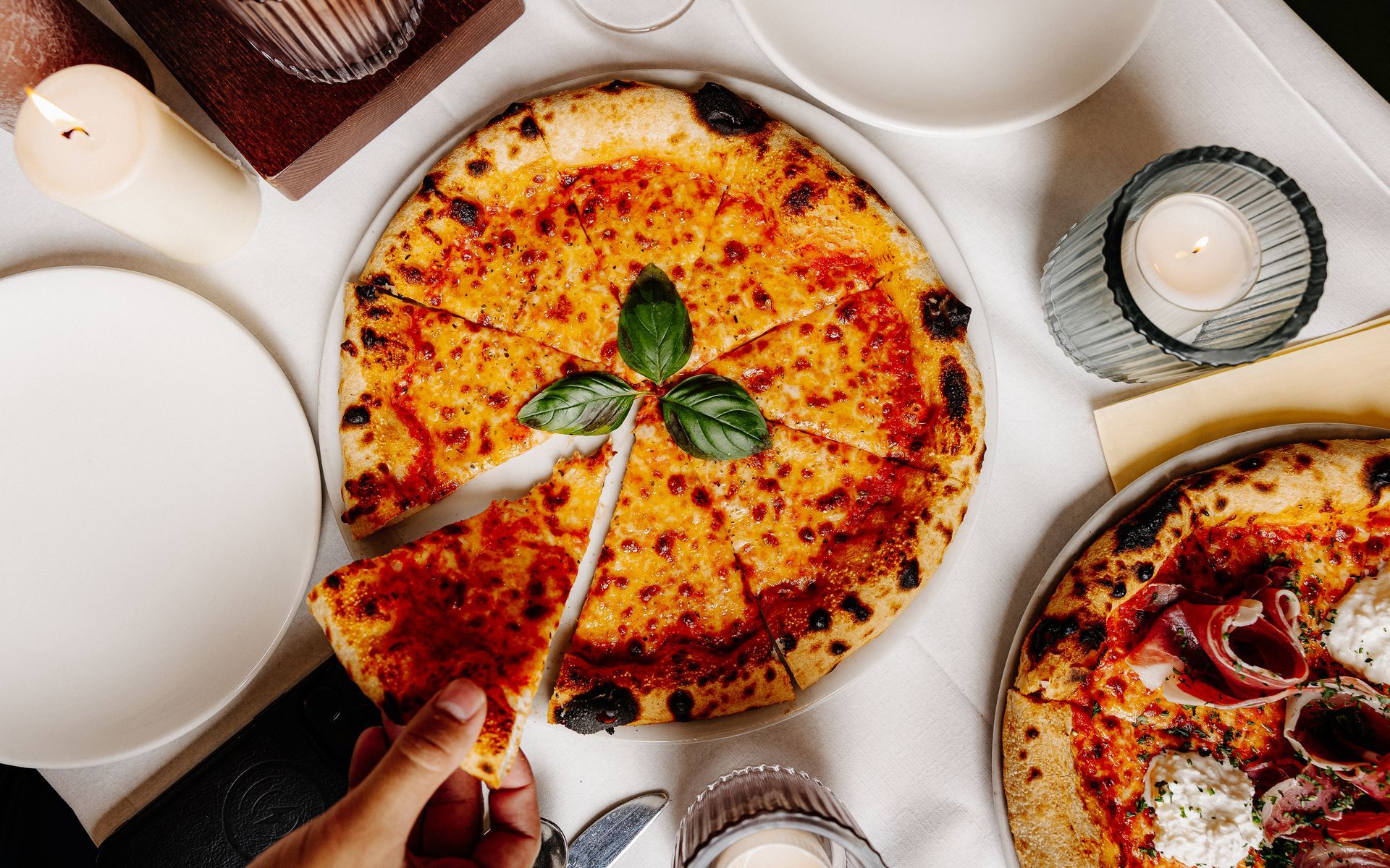 Close-up van twee pizza's op een tafel, waarvan er één wordt gesneden. Ook kaarsen en borden zijn aanwezig. Bij L'Essenza Rotterdam.