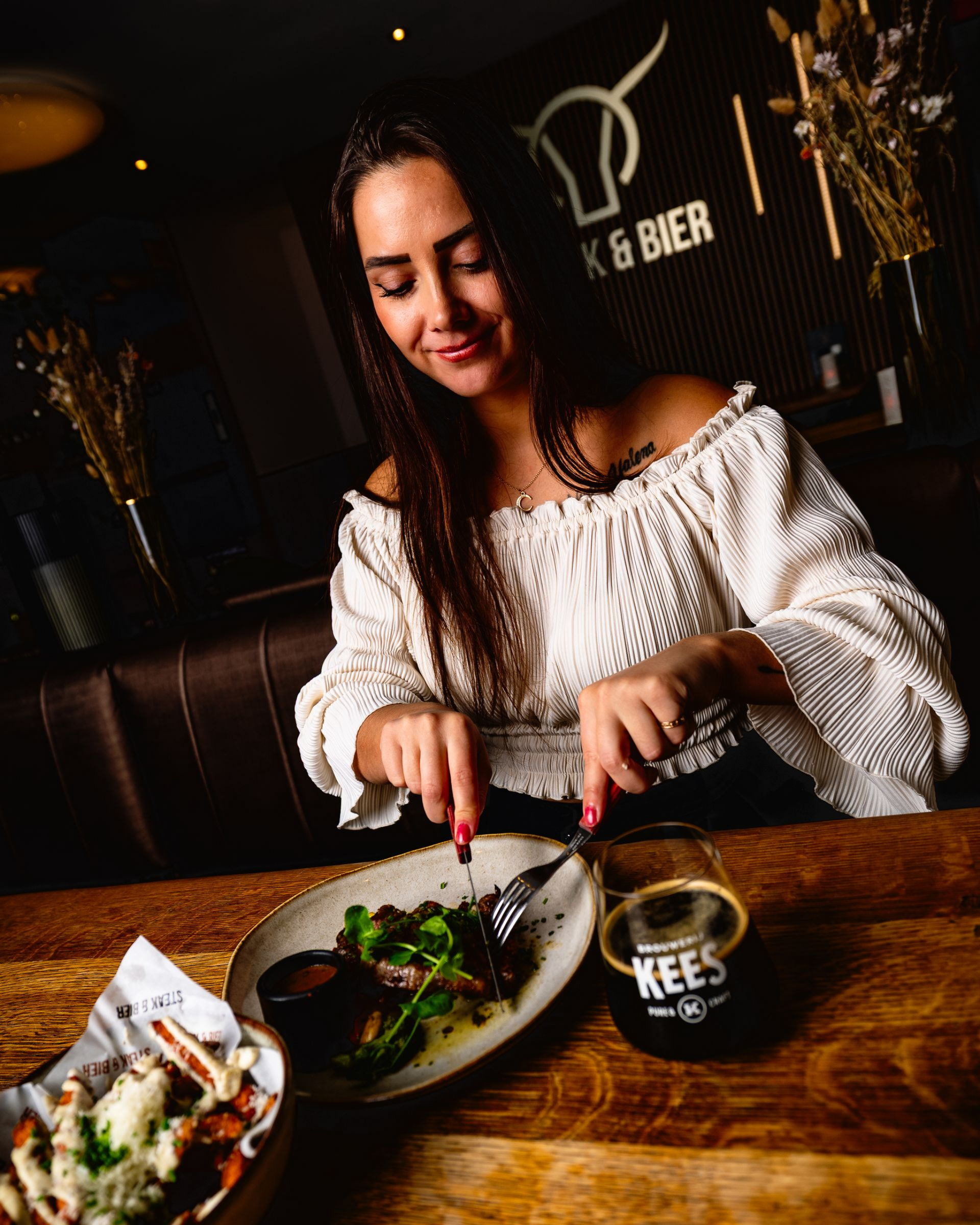 Vrouw eet in een restaurant, met mes en vork in de hand, vlakbij een bord met eten en een biertje.