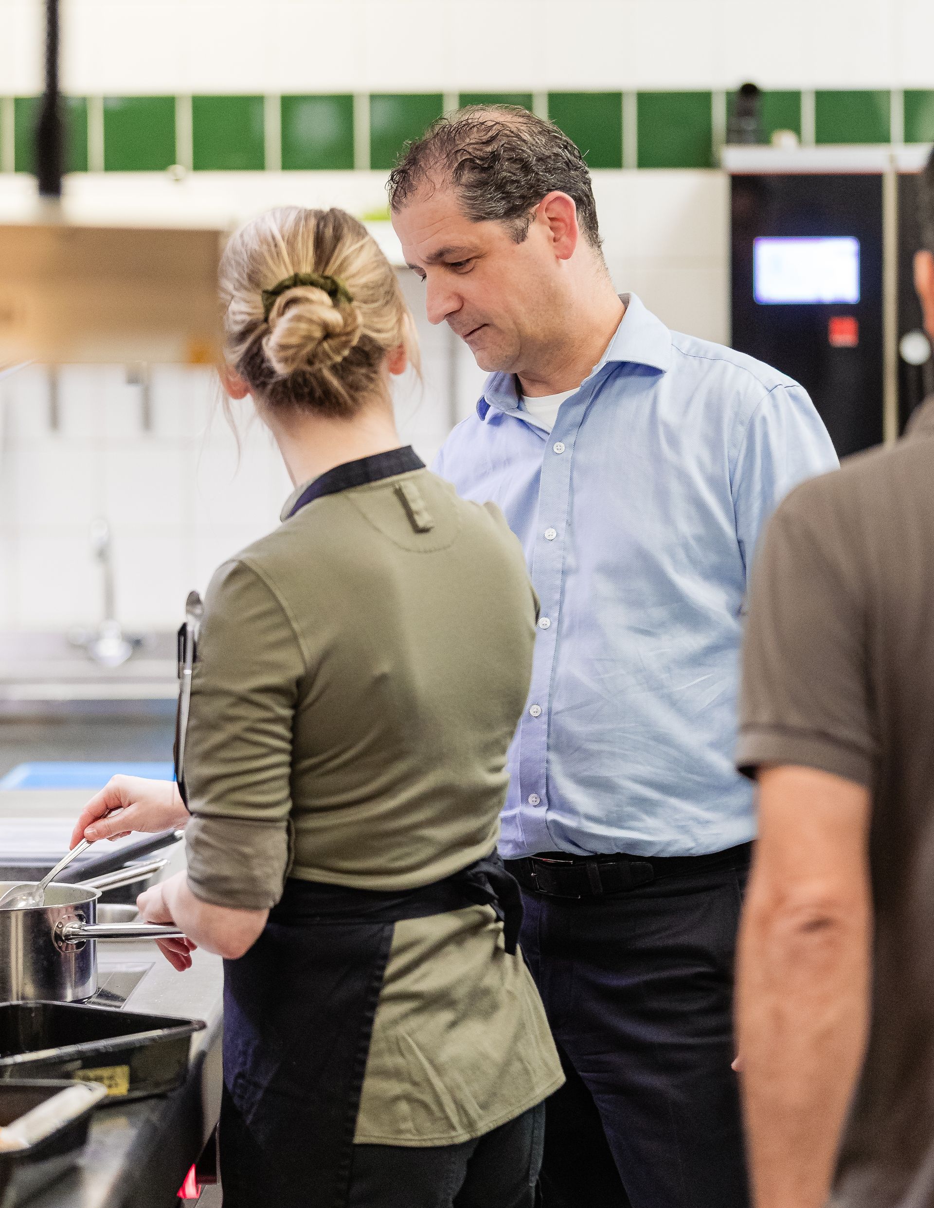 Een man in een blauw shirt overlegt met iemand die een schort draagt in een keuken over de bereiding van voedsel.