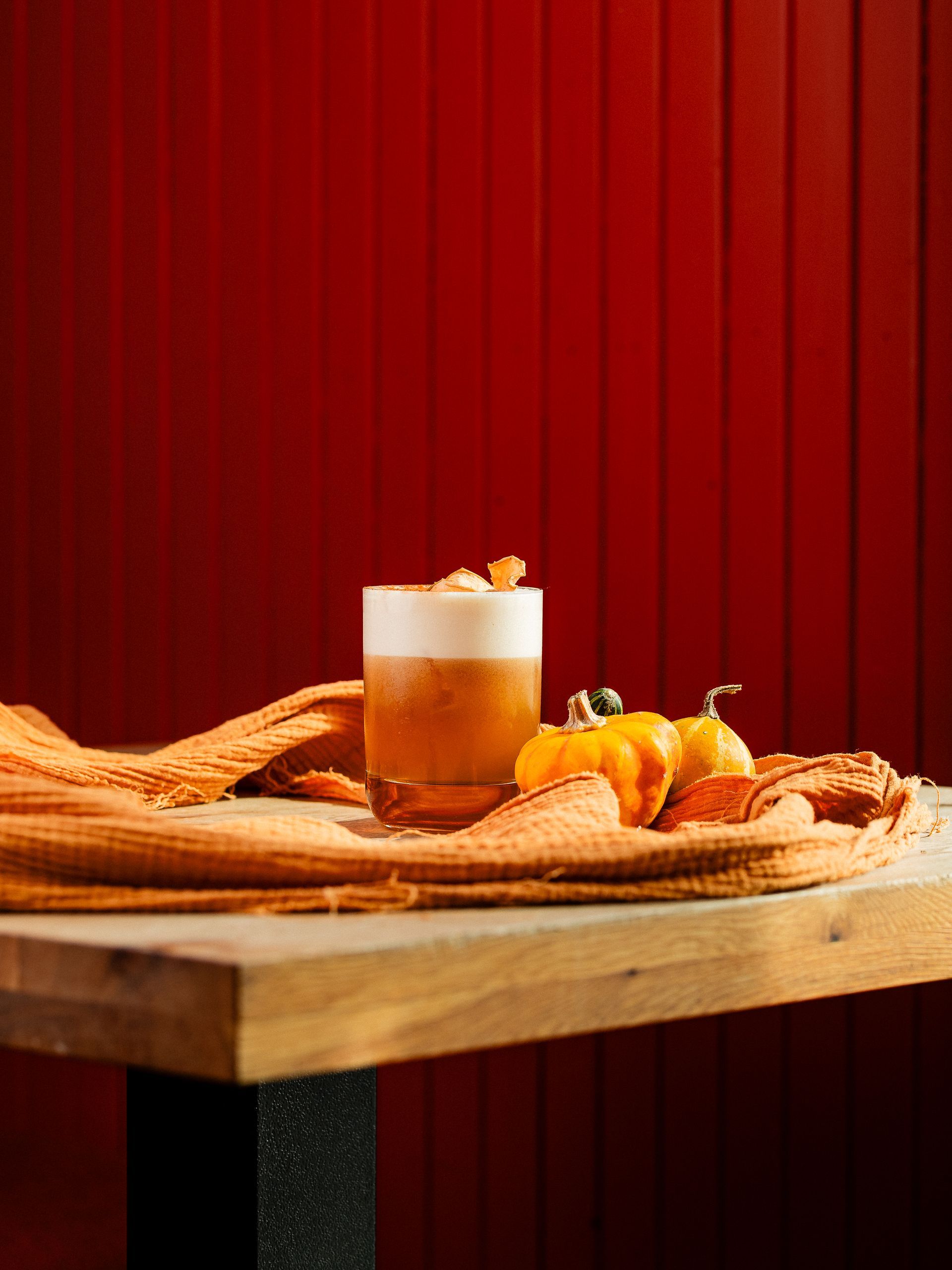 Een cocktail in een glas, afgetopt met schuim, naast mini-pompoenen op een oranje tafelkleed, op een houten tafel met een rode achtergrond.