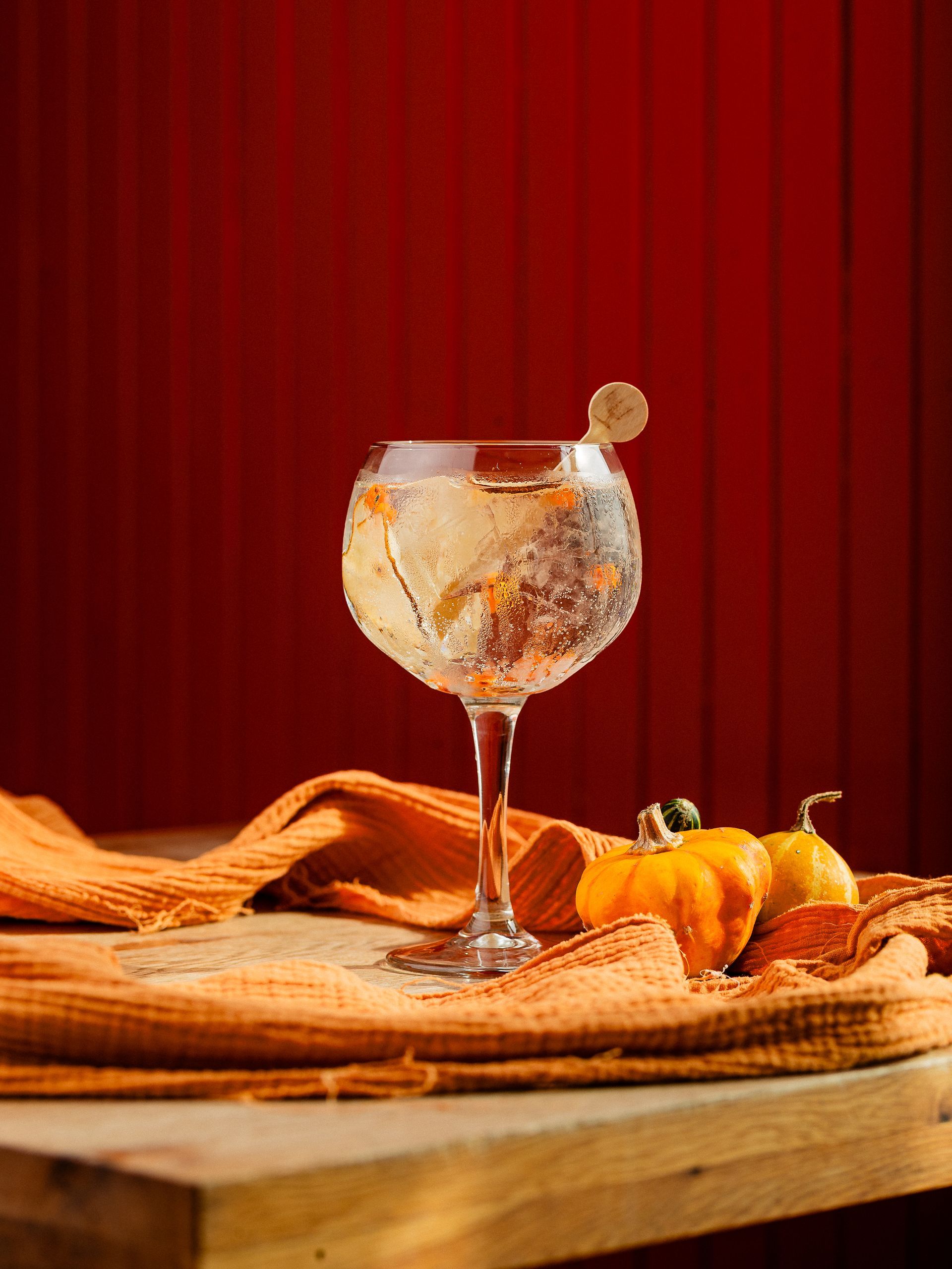 Cocktail in een wijnglas met drijvende stukjes; pompoenen en een oranje tafelkleed op een houten tafel. Rode achtergrond.