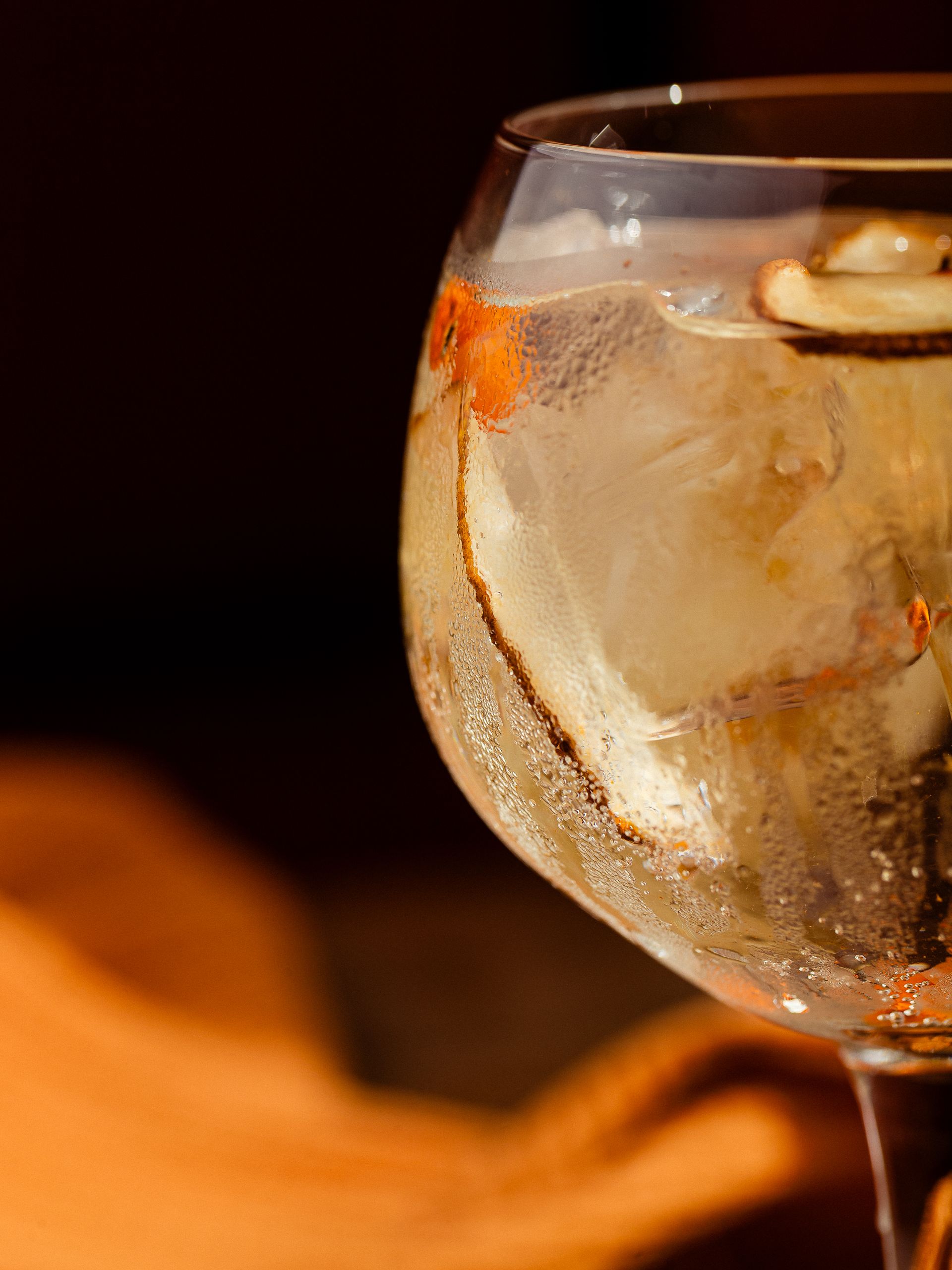 Close-up van een heldere cocktail in een glas op voet met ijs, bubbels en garnering, badend in warm licht.
